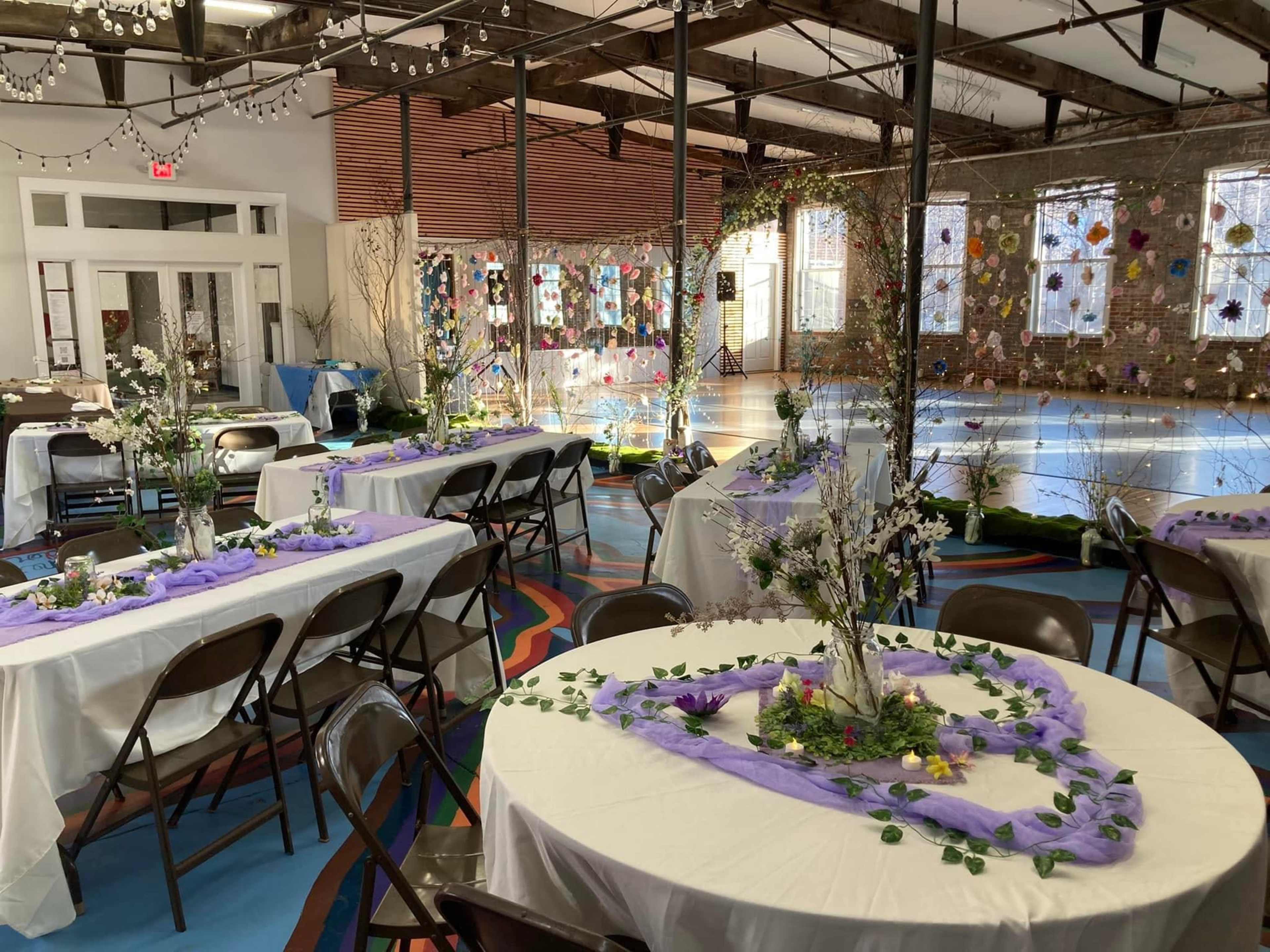 A spacious event hall features several round tables adorned with floral centerpieces, surrounded by chairs and decorated with fabric drapes, while colorful artwork adorns the walls.