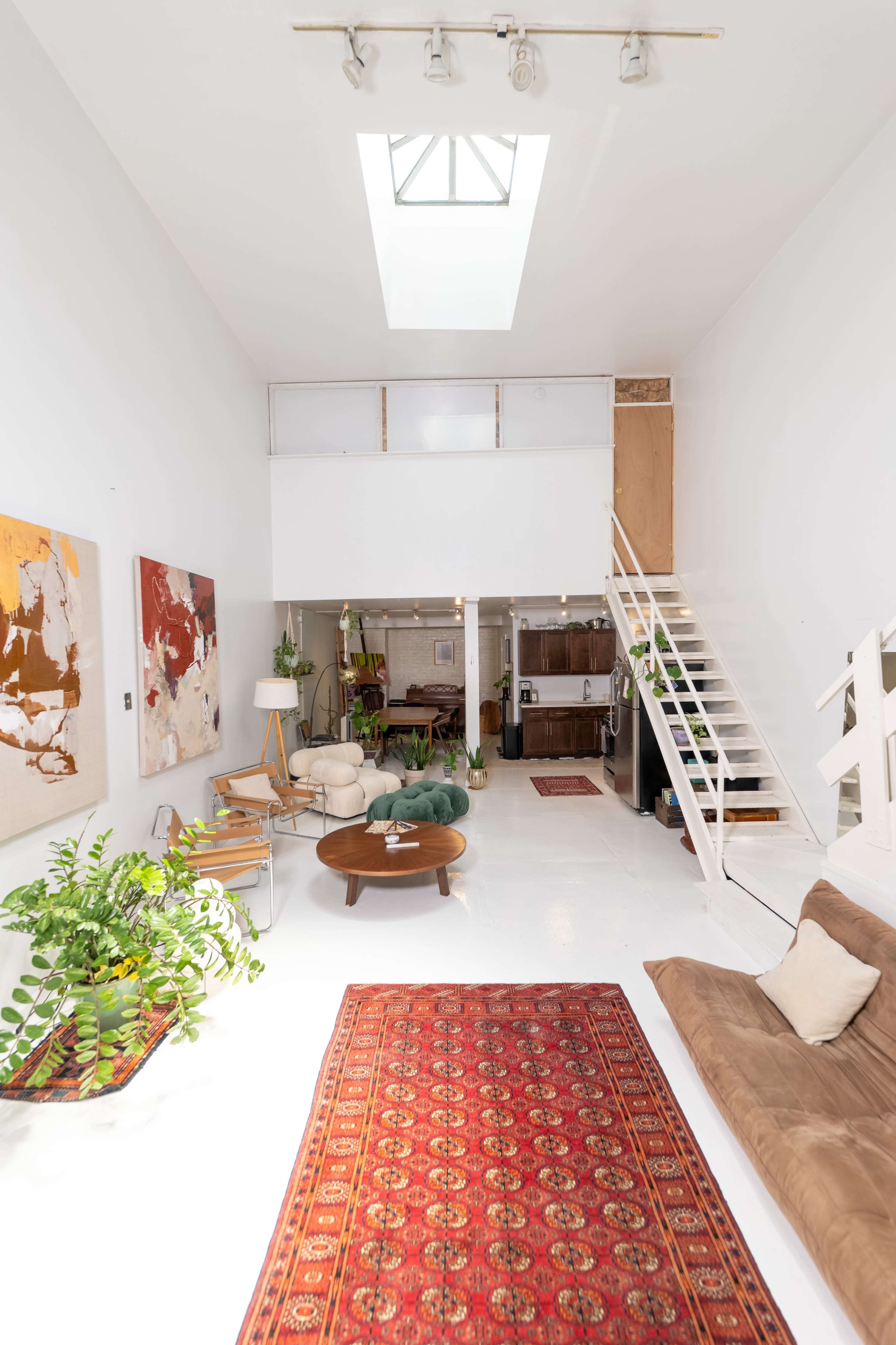 A spacious room with white walls, a skylight above, a patterned rug on a polished floor, and a staircase leading to an upper level.