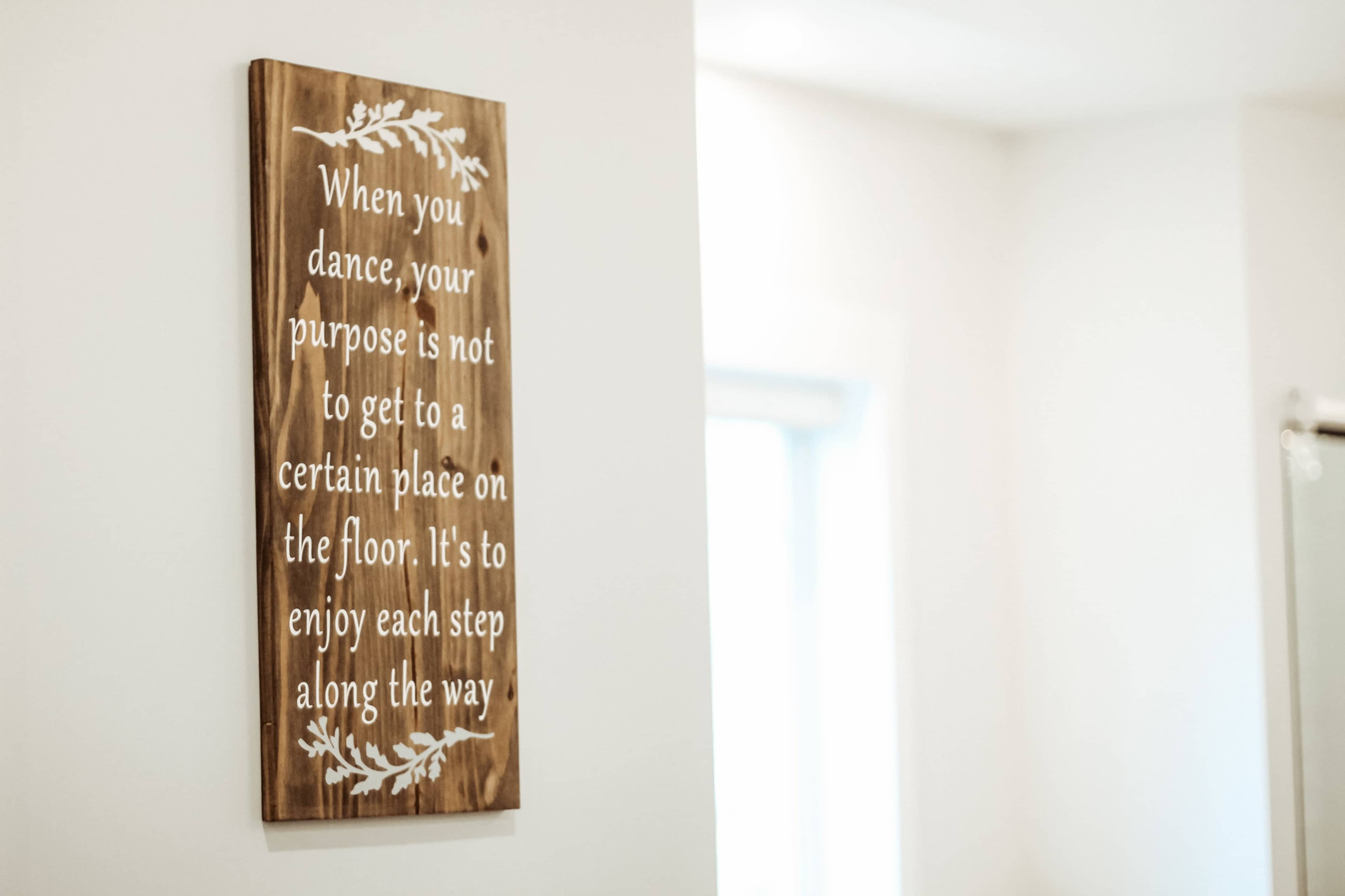 A wooden sign featuring white text with a motivational message about dancing is mounted on a light-colored wall.