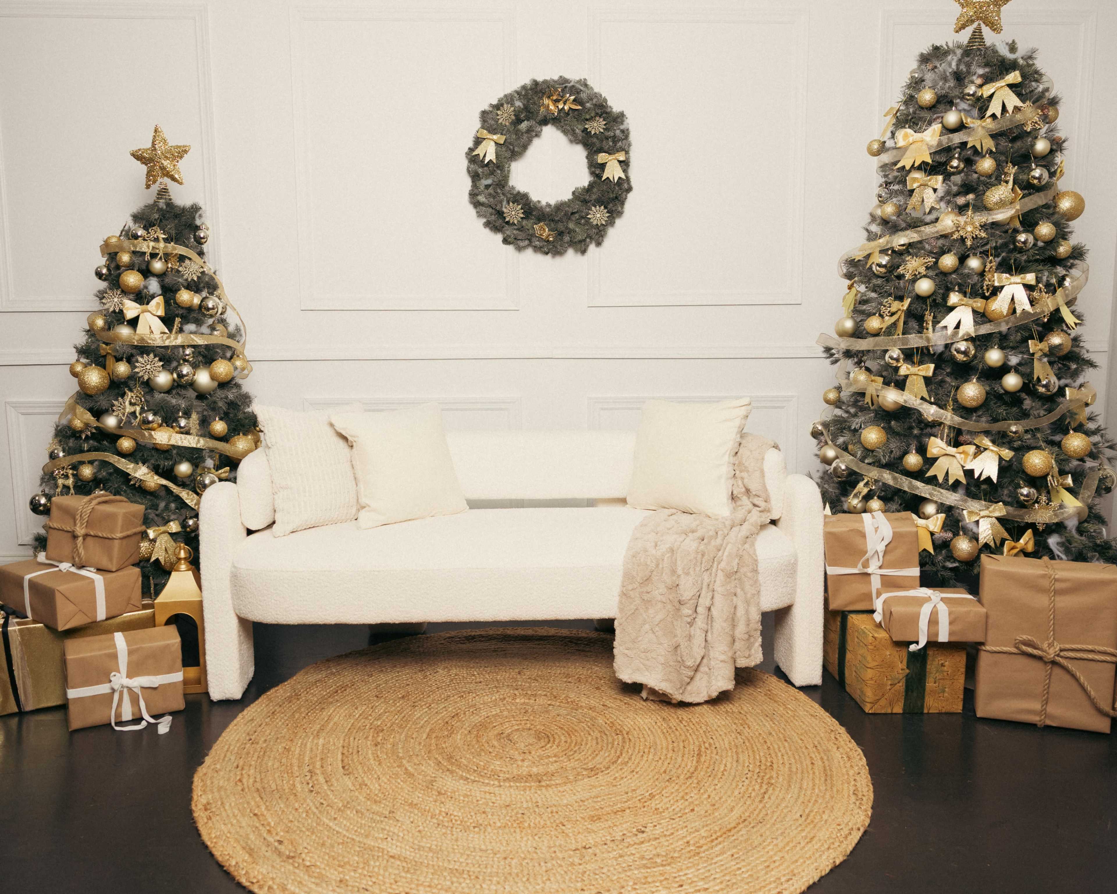 A cozy living room features two decorated Christmas trees, a white sofa with cushions, a round jute rug, and several gift boxes arranged around the space.