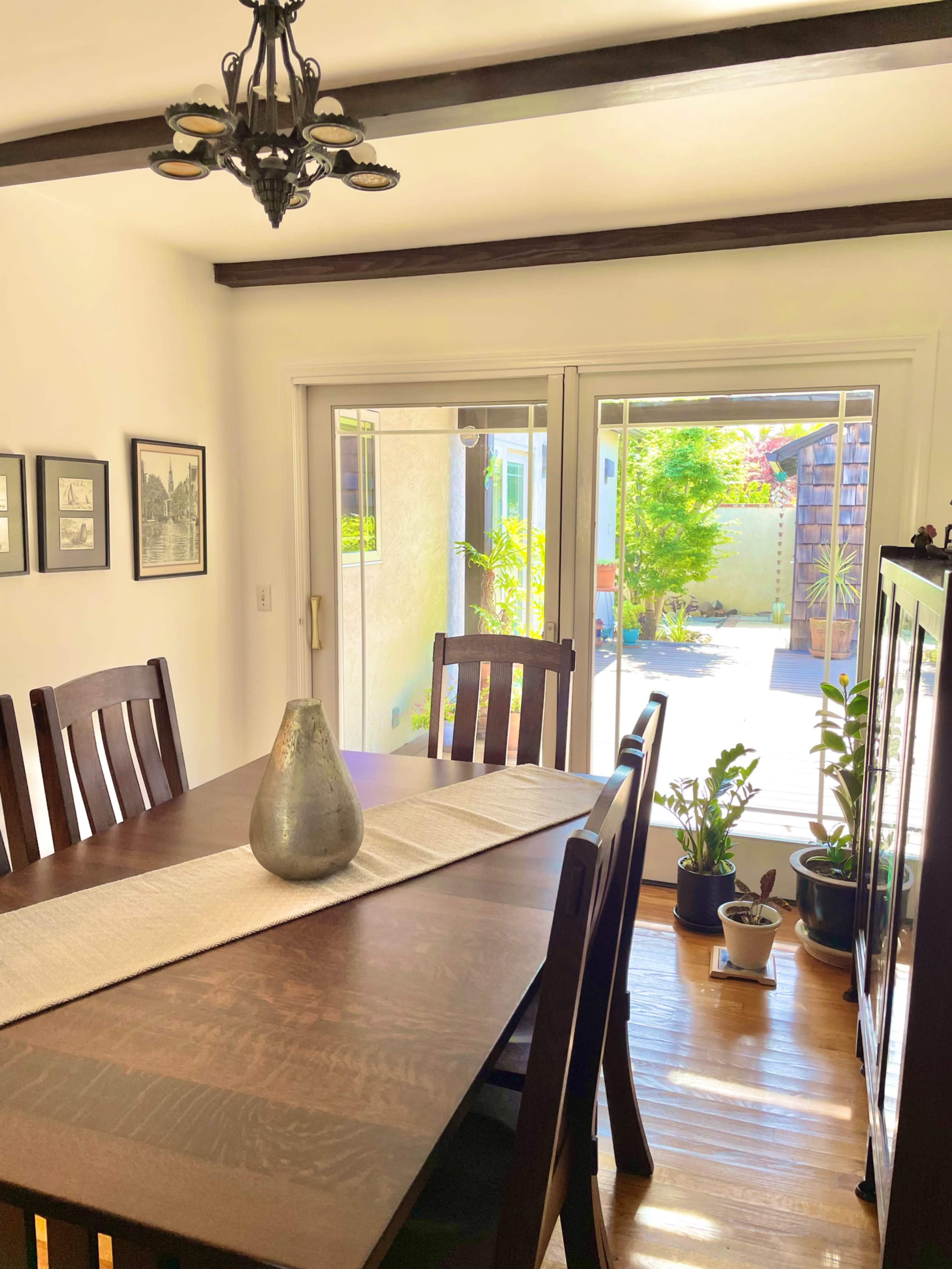 A dining area features a wooden table with chairs, a centerpiece vase, and large sliding doors opening to a yard with plants.