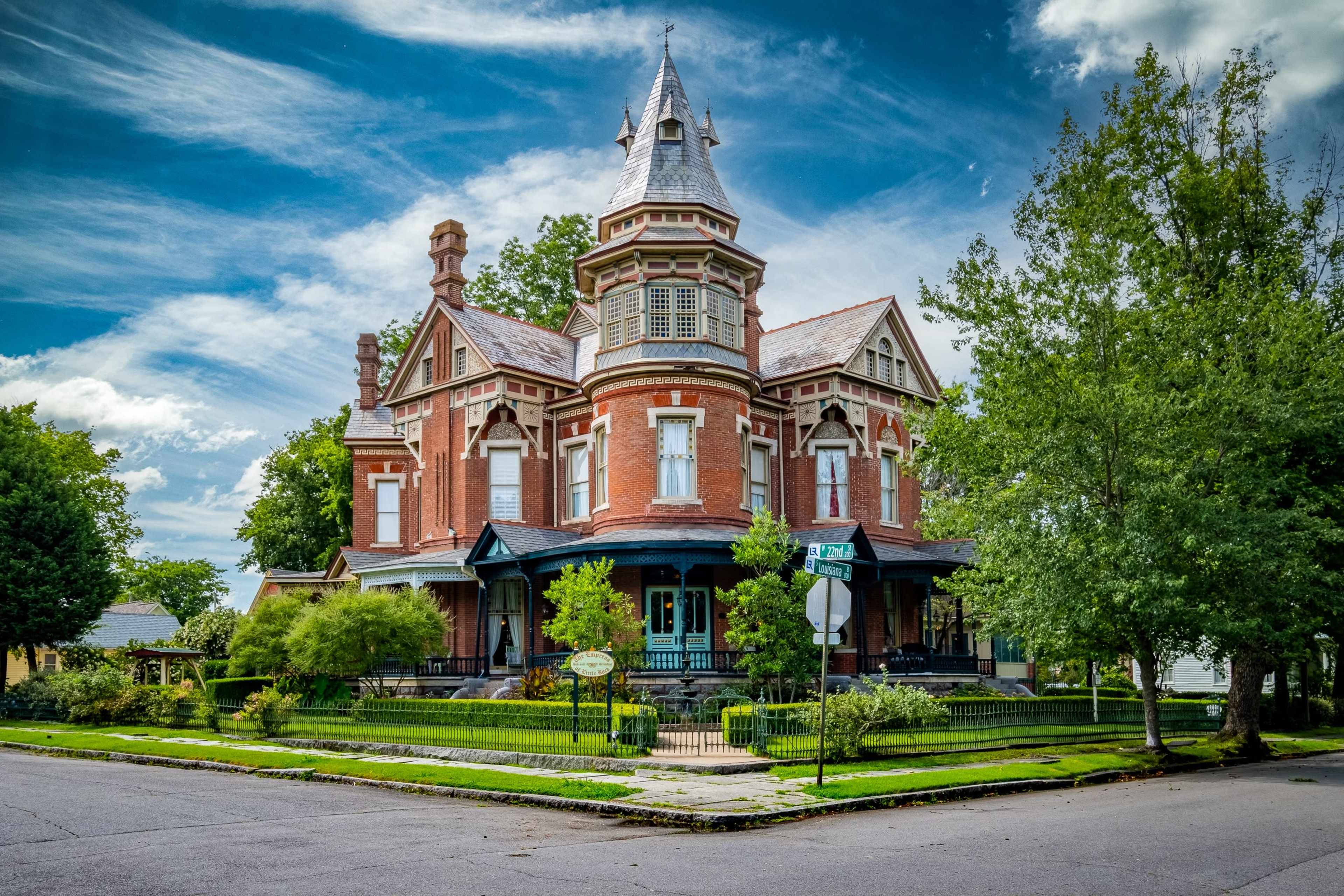 The image shows a large, three-story Victorian house with elaborate architectural details, a tower, and a well-maintained garden at the corner of a street.