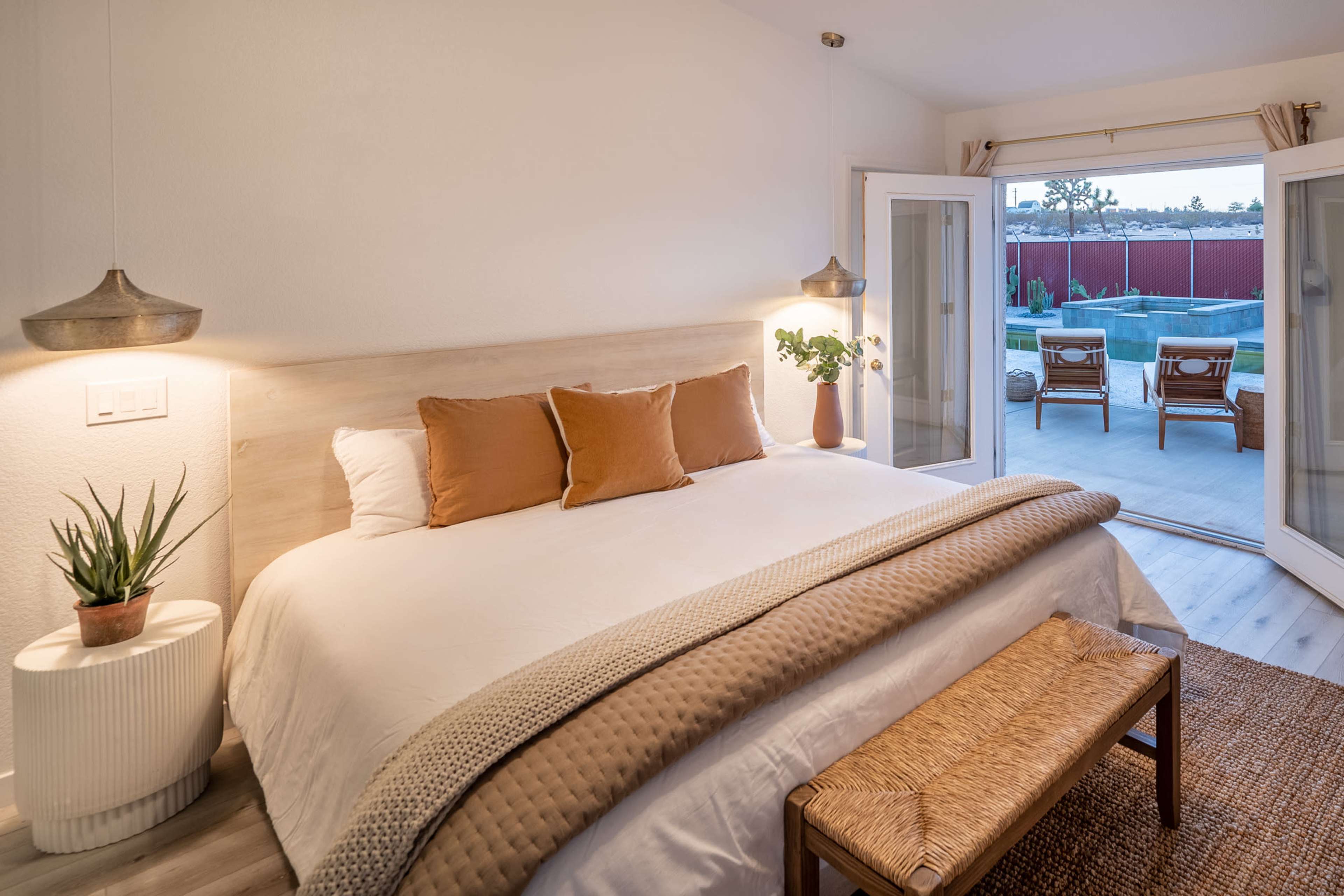 The image shows a modern bedroom featuring a large bed with decorative pillows, a bench at the foot, and sliding glass doors leading to an outdoor area with chairs.