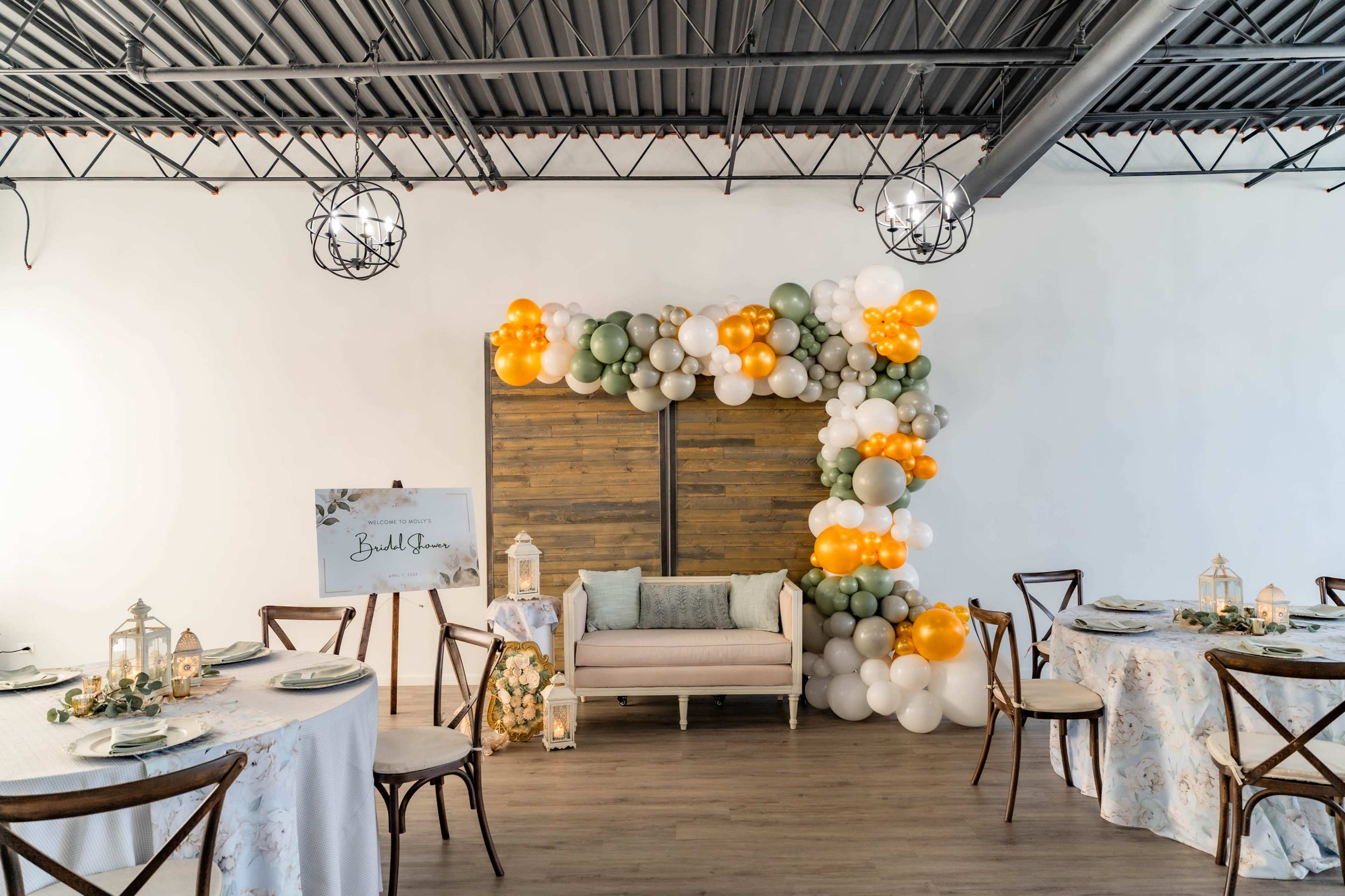 A decorated event space with balloon arrangements in orange, green, and white, a wooden backdrop, and elegantly set tables.