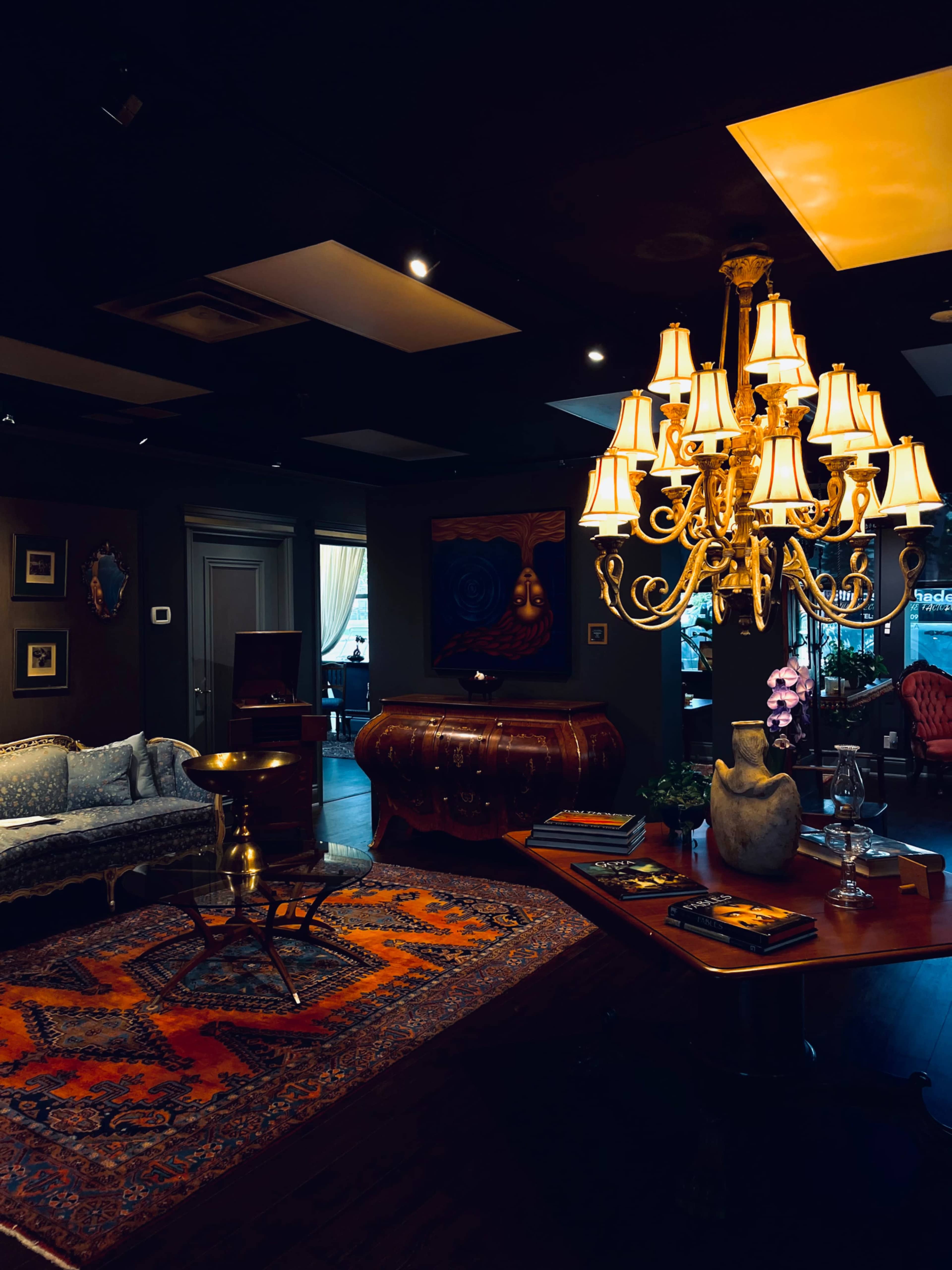 A stylish, dimly lit room features a vintage sofa, a decorative chandelier, and ornate furnishings arranged around a colorful area rug.
