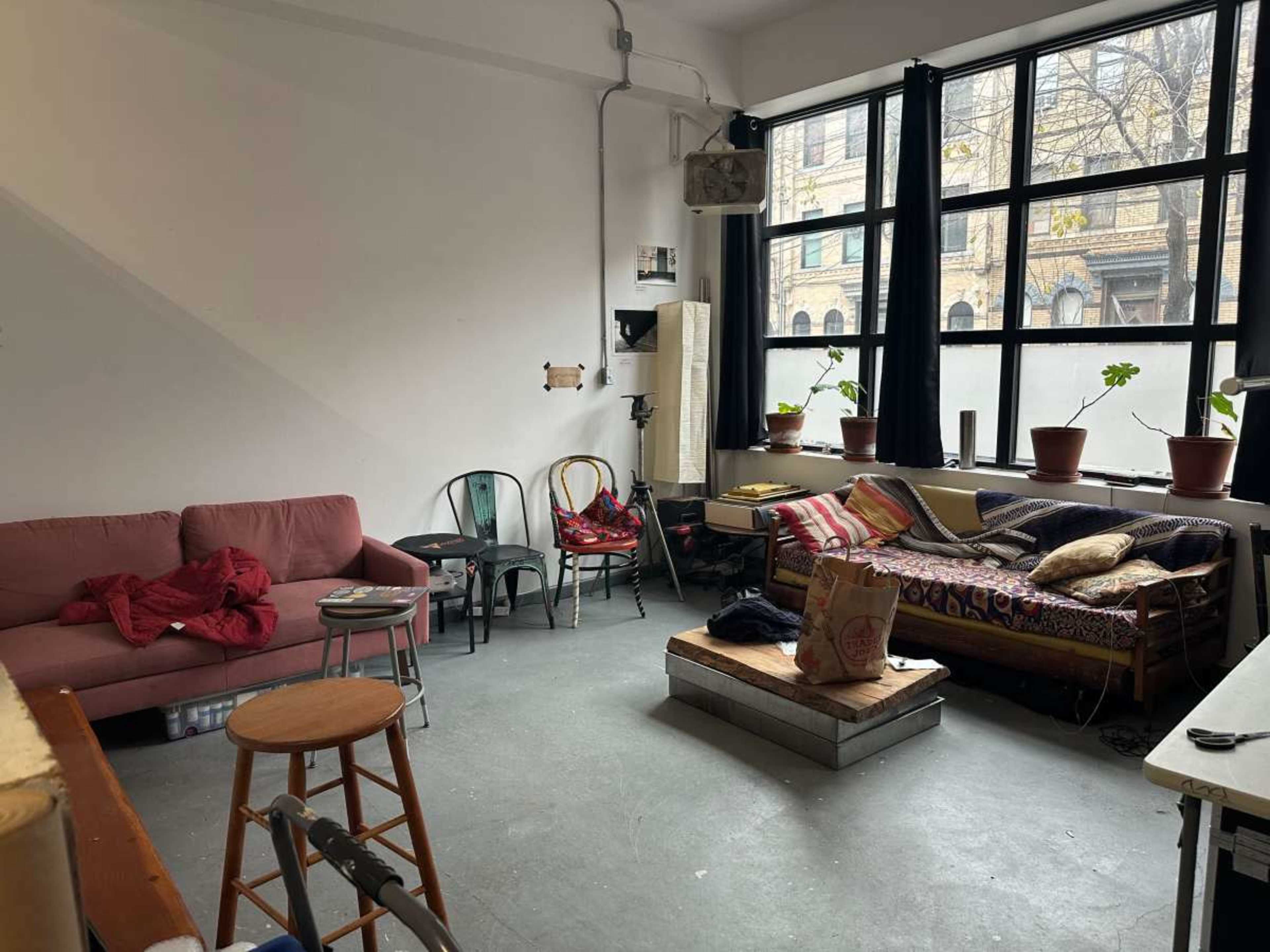 The image shows a partially furnished room with a pink couch, a wooden stool, a patterned sofa, potted plants on windowsills, and a table in the corner.