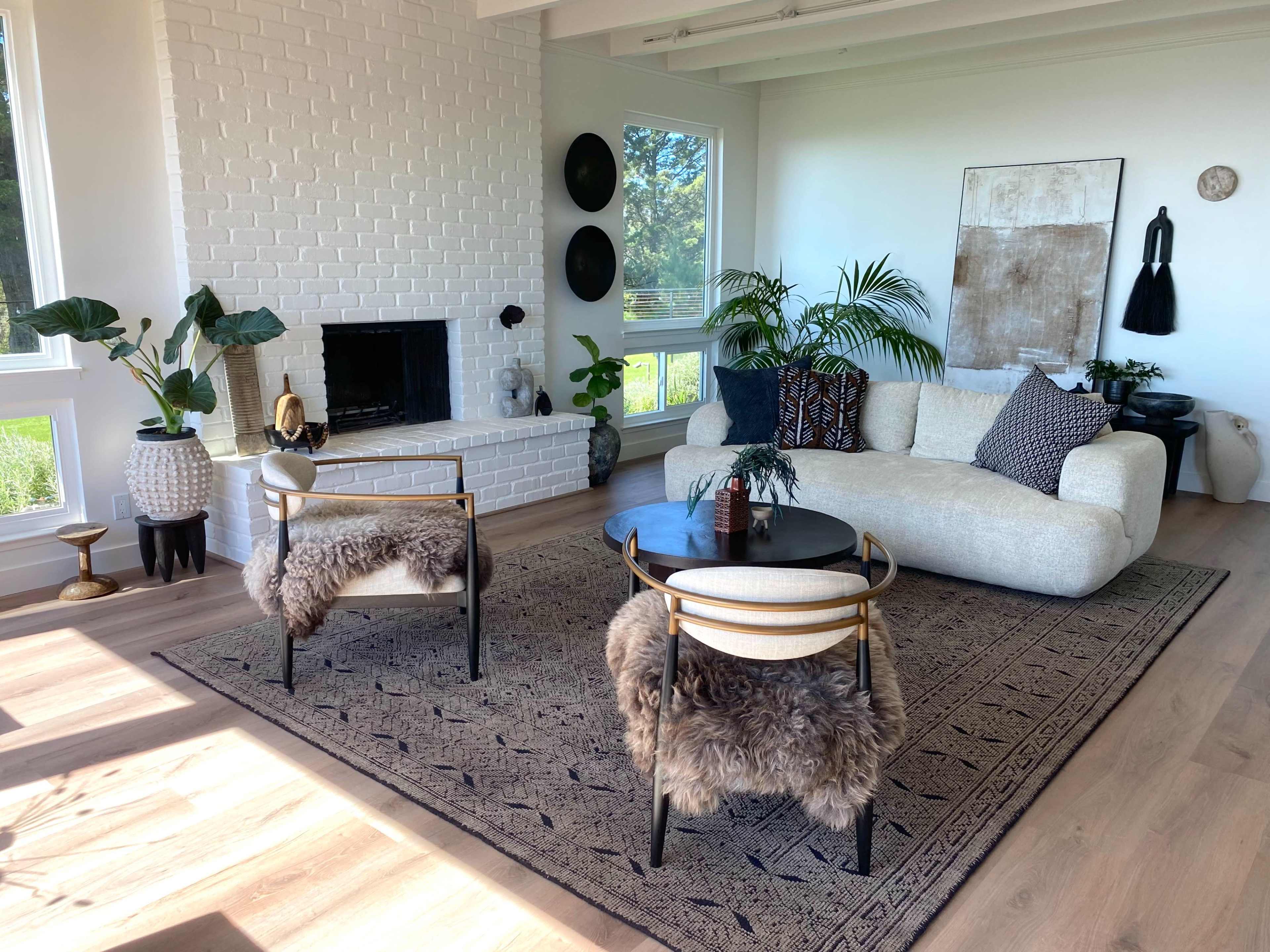 The image shows a modern living room with a white brick fireplace, two chairs with fur throws, a curved sofa, and large windows allowing natural light to fill the space.