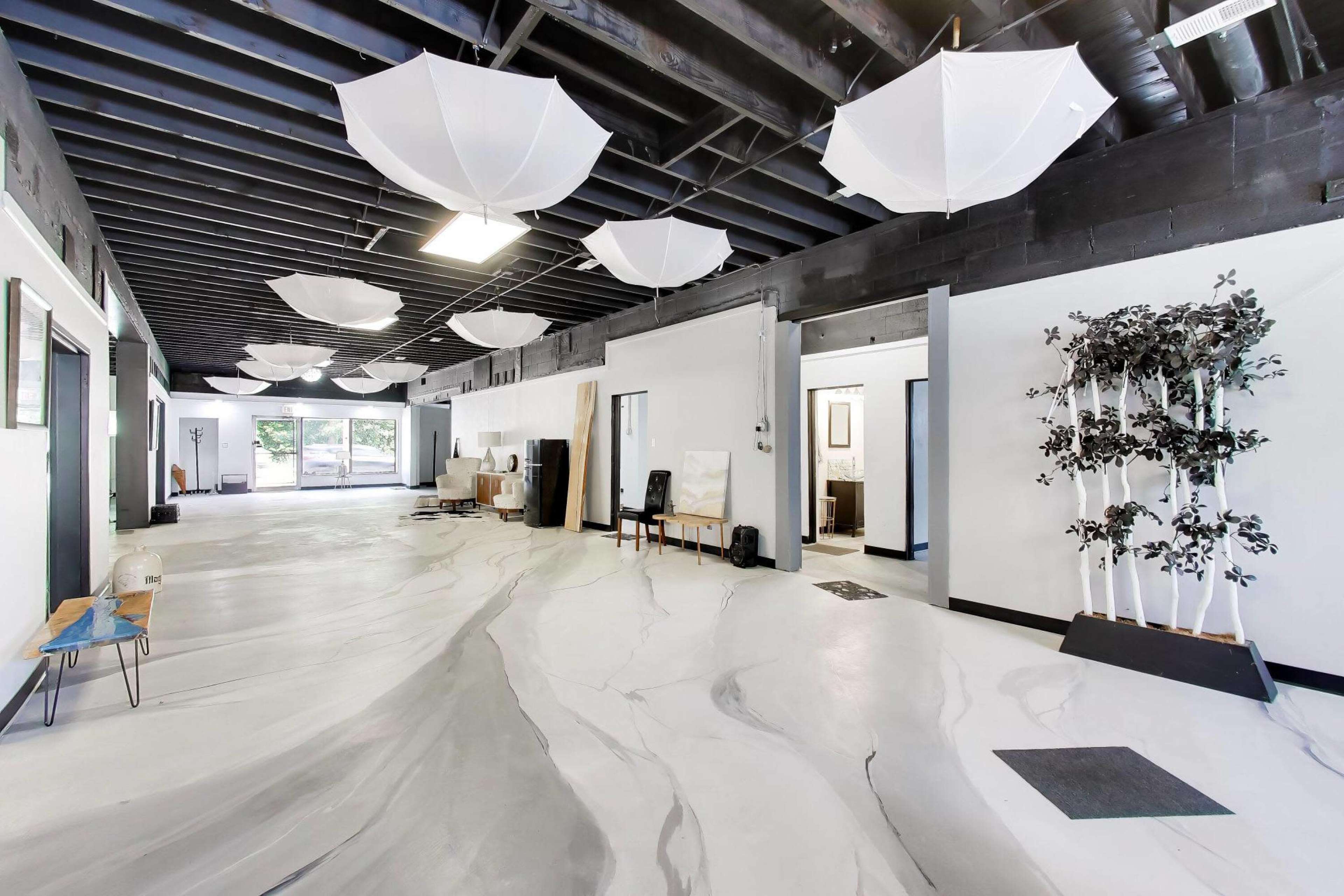 A spacious, well-lit interior features a white floor with gray marbling, adorned with suspended umbrellas and minimalist decor.
