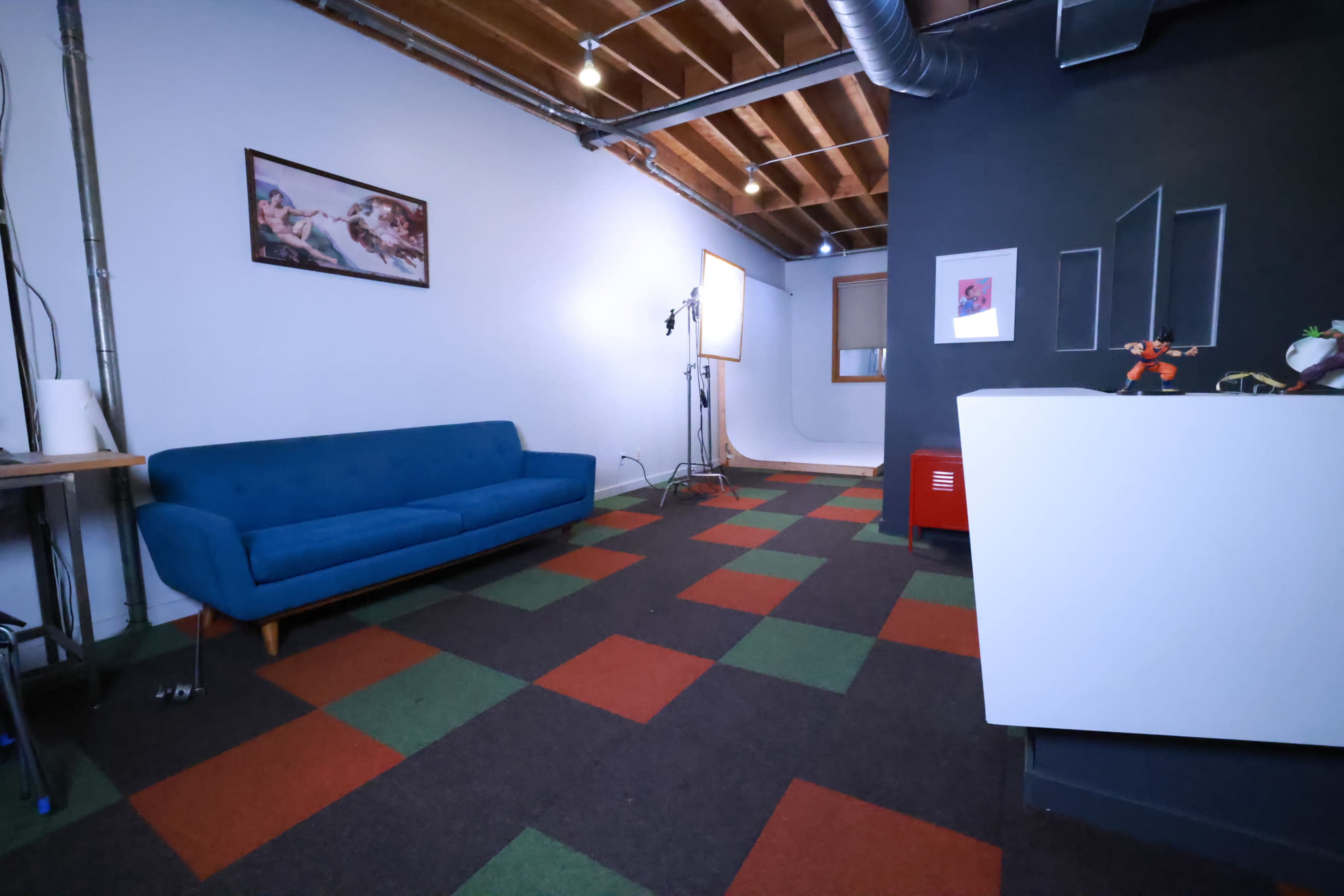 The image shows a spacious room with a blue couch, a framed artwork on the wall, a lighting setup, and a multicolored carpet.