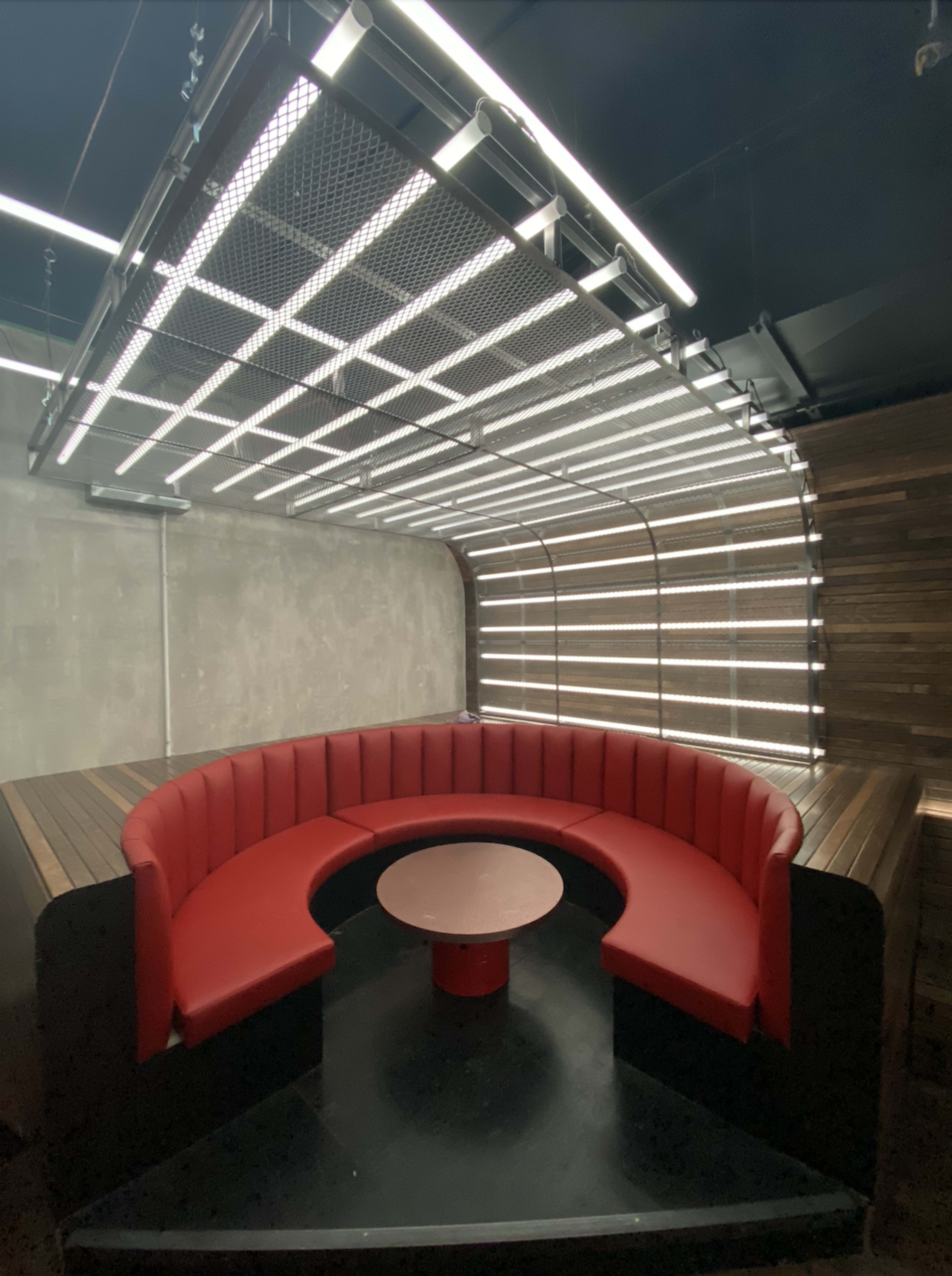 The image shows a modern round seating area with red upholstery and a circular table, illuminated by overhead linear lighting against a textured wall.