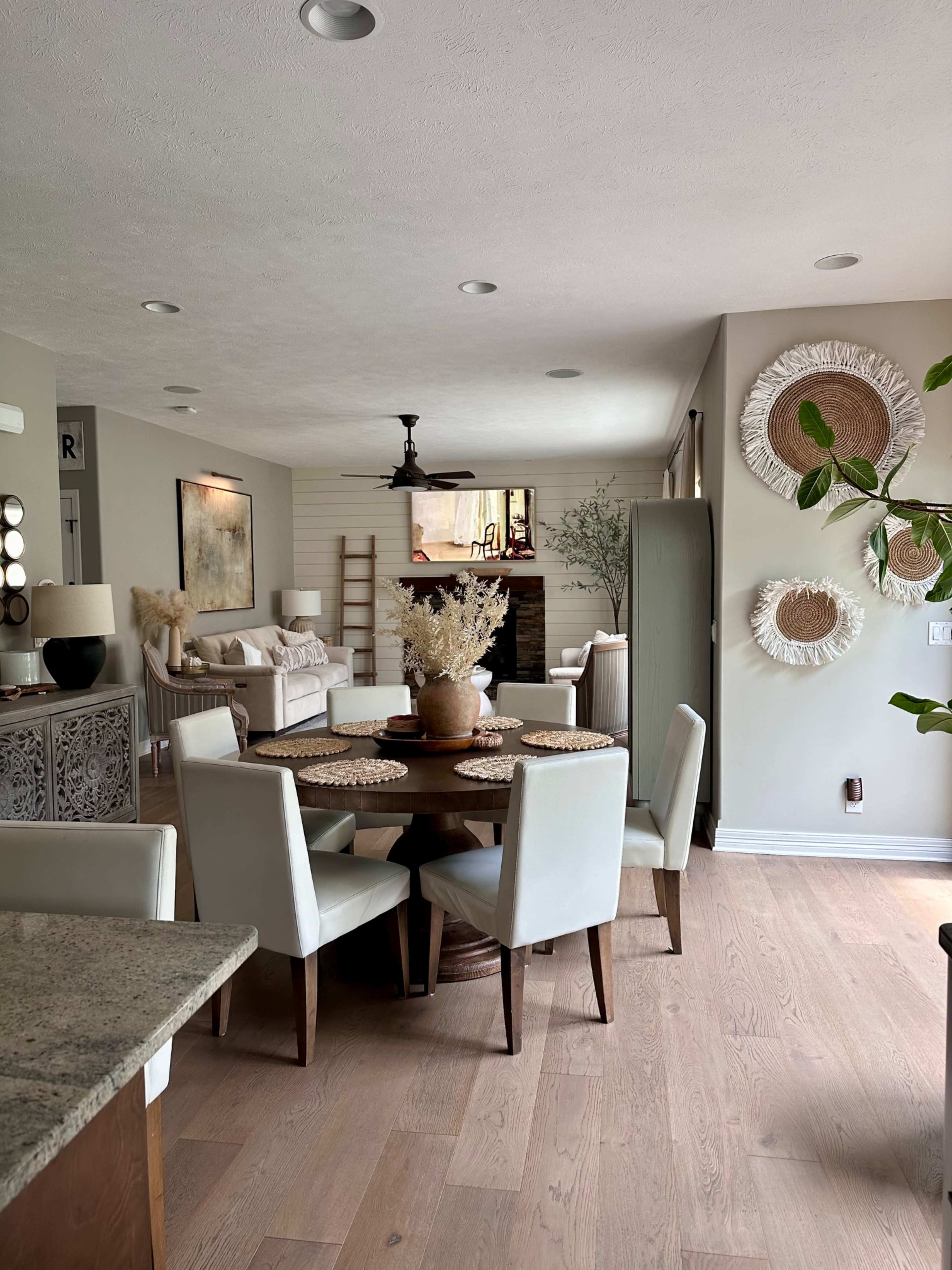 The image shows a modern living and dining area featuring a round wooden table with white chairs, a beige sofa, and decorative wall accents.