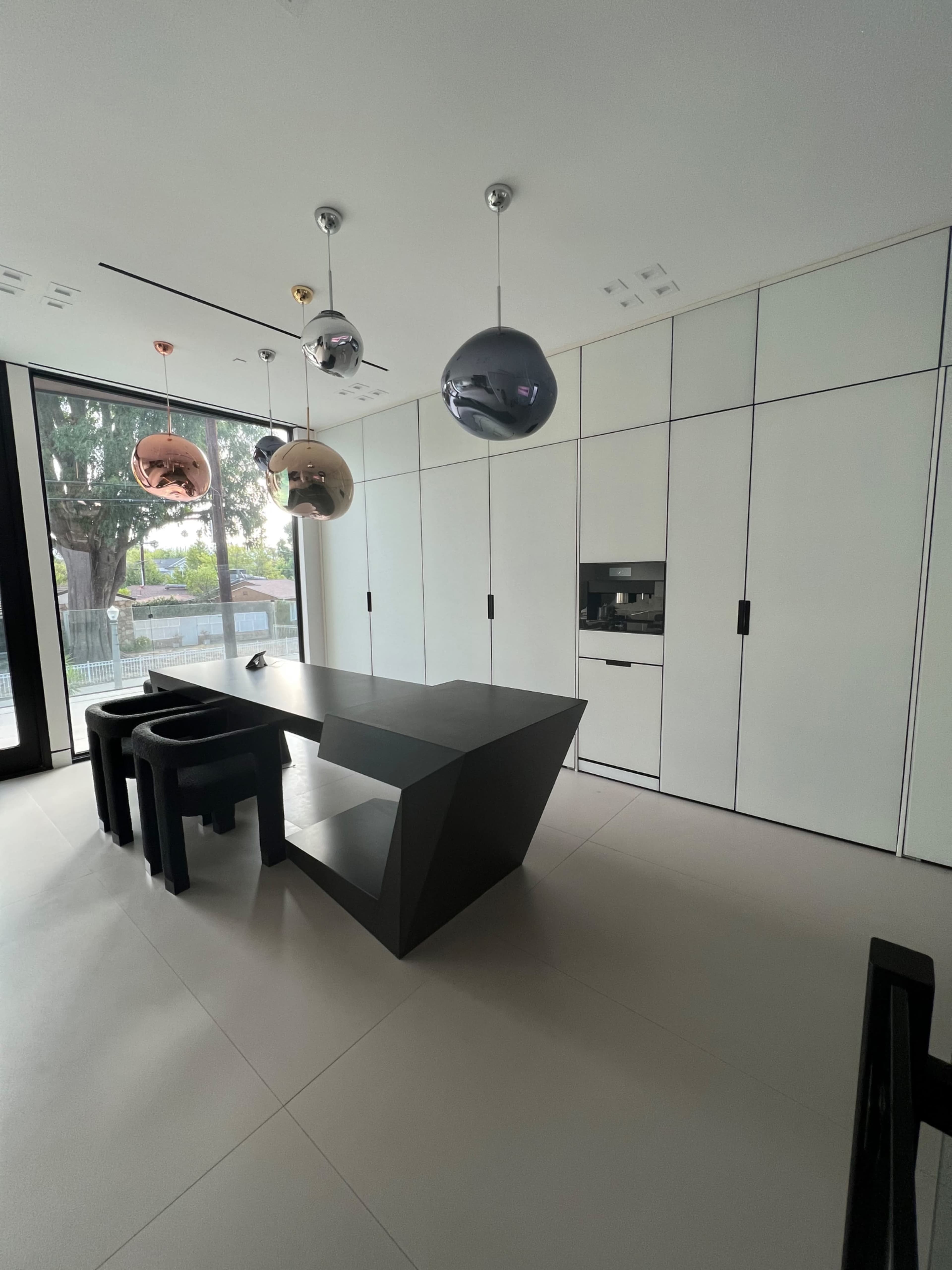 A modern kitchen with a black geometric dining table, black stools, and several pendant lights hanging from the ceiling.