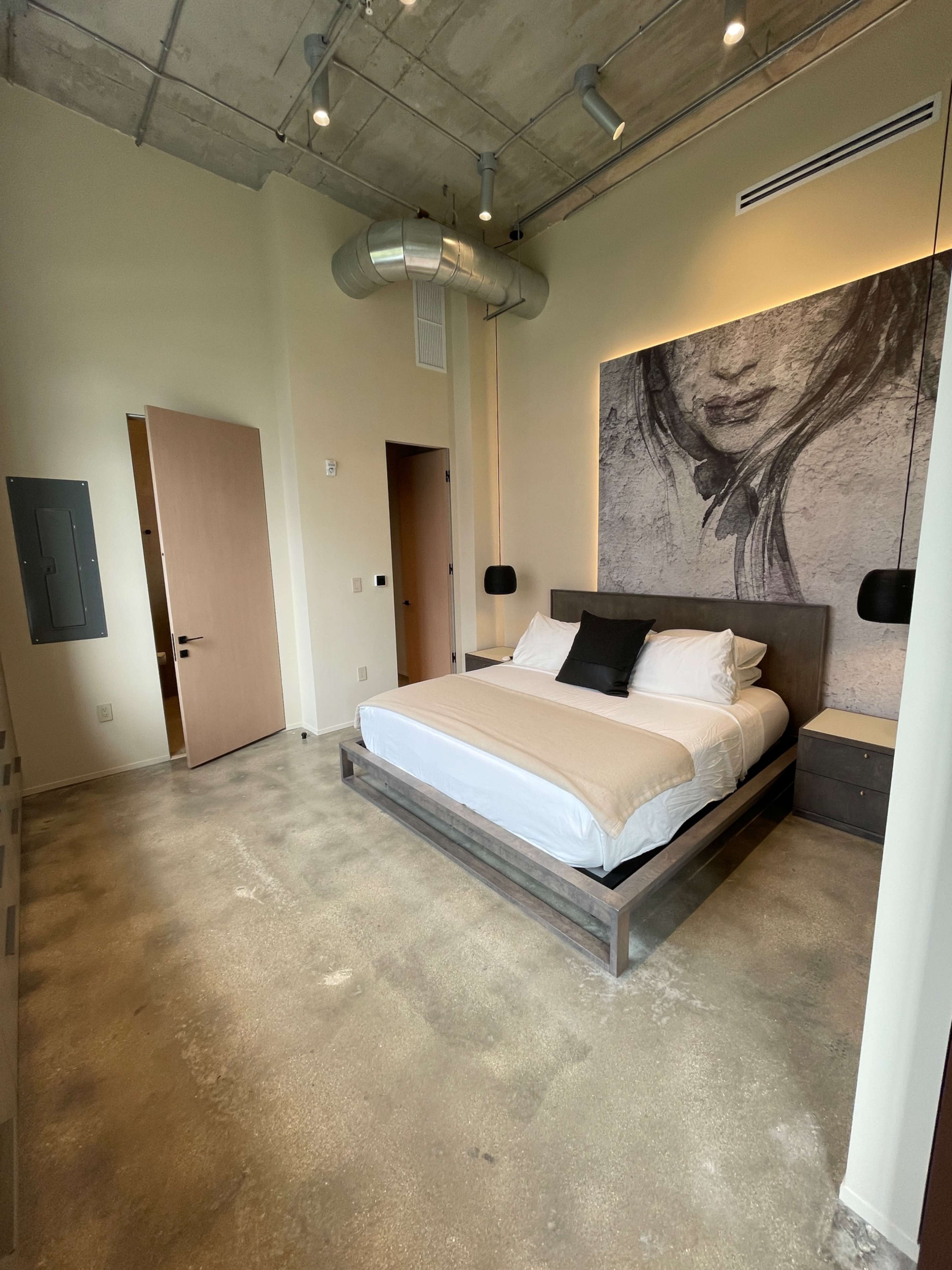 A minimalistic bedroom with a large bed, a prominent wall mural, and a sleek concrete floor.