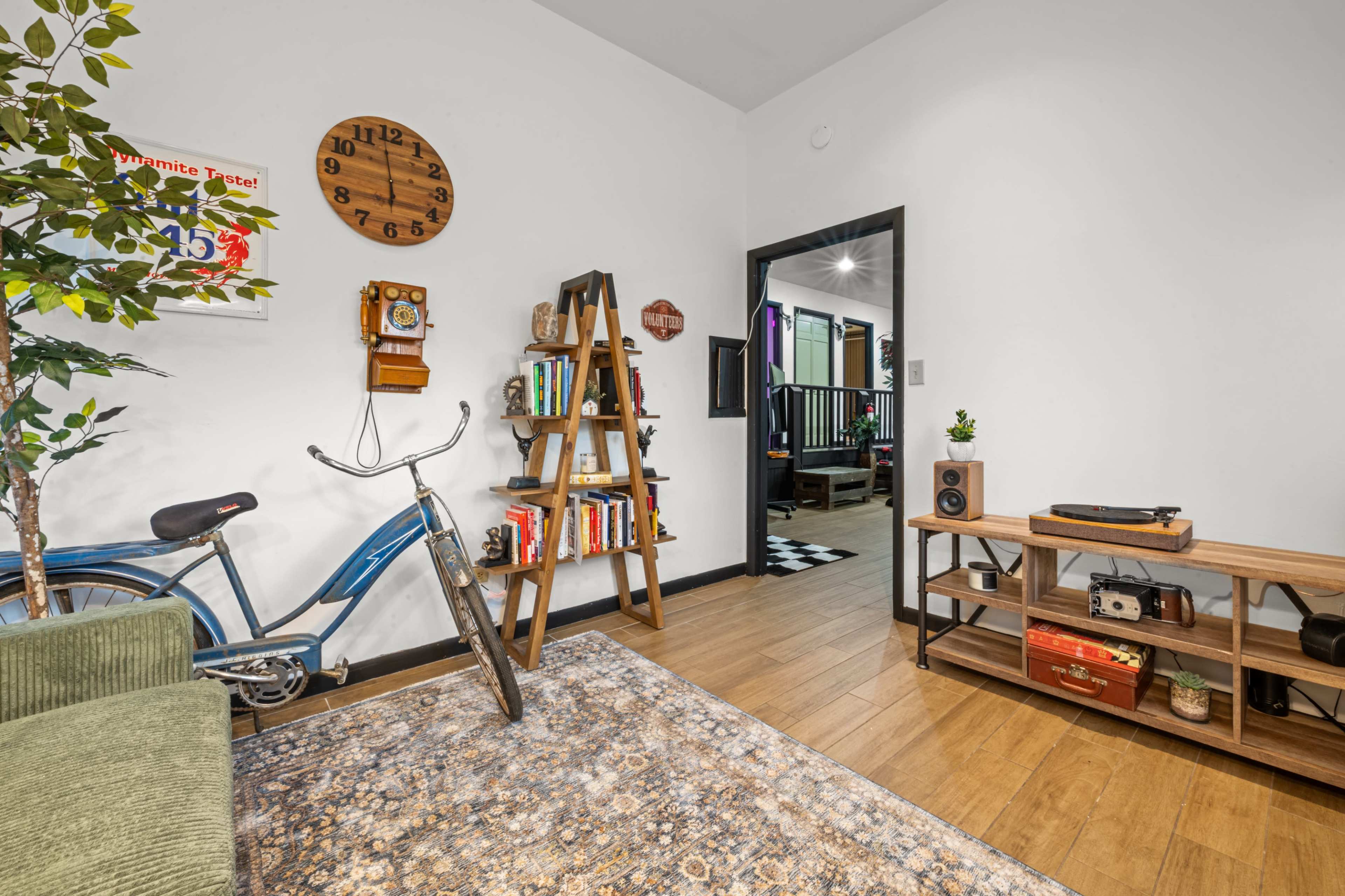 The image shows a cozy room featuring a vintage bicycle, a wooden bookshelf filled with books, and a record player on a side table, with a doorway leading to another area.