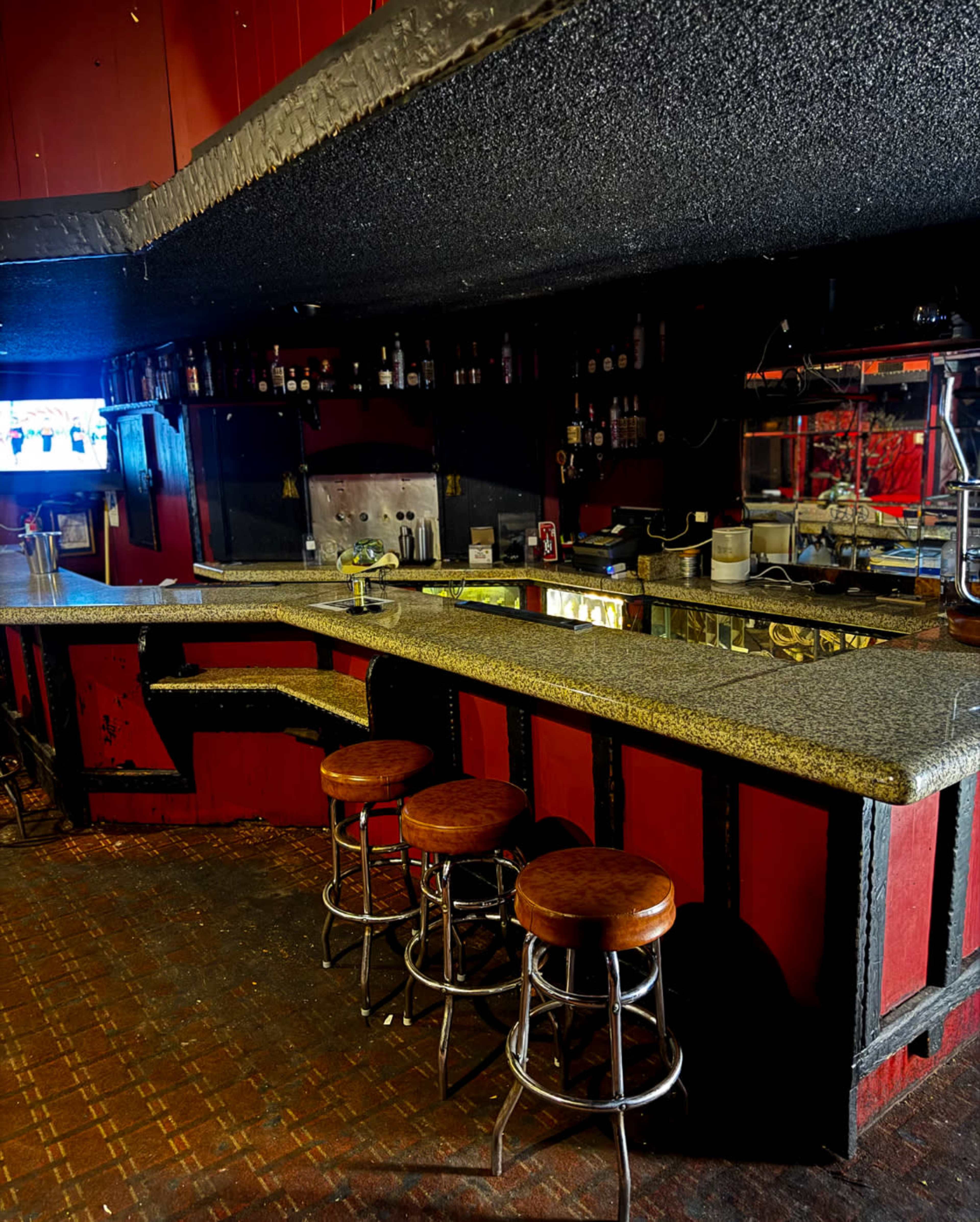 A dimly lit bar interior features a granite countertop with three metal stools, surrounded by red walls and shelves lined with bottles.