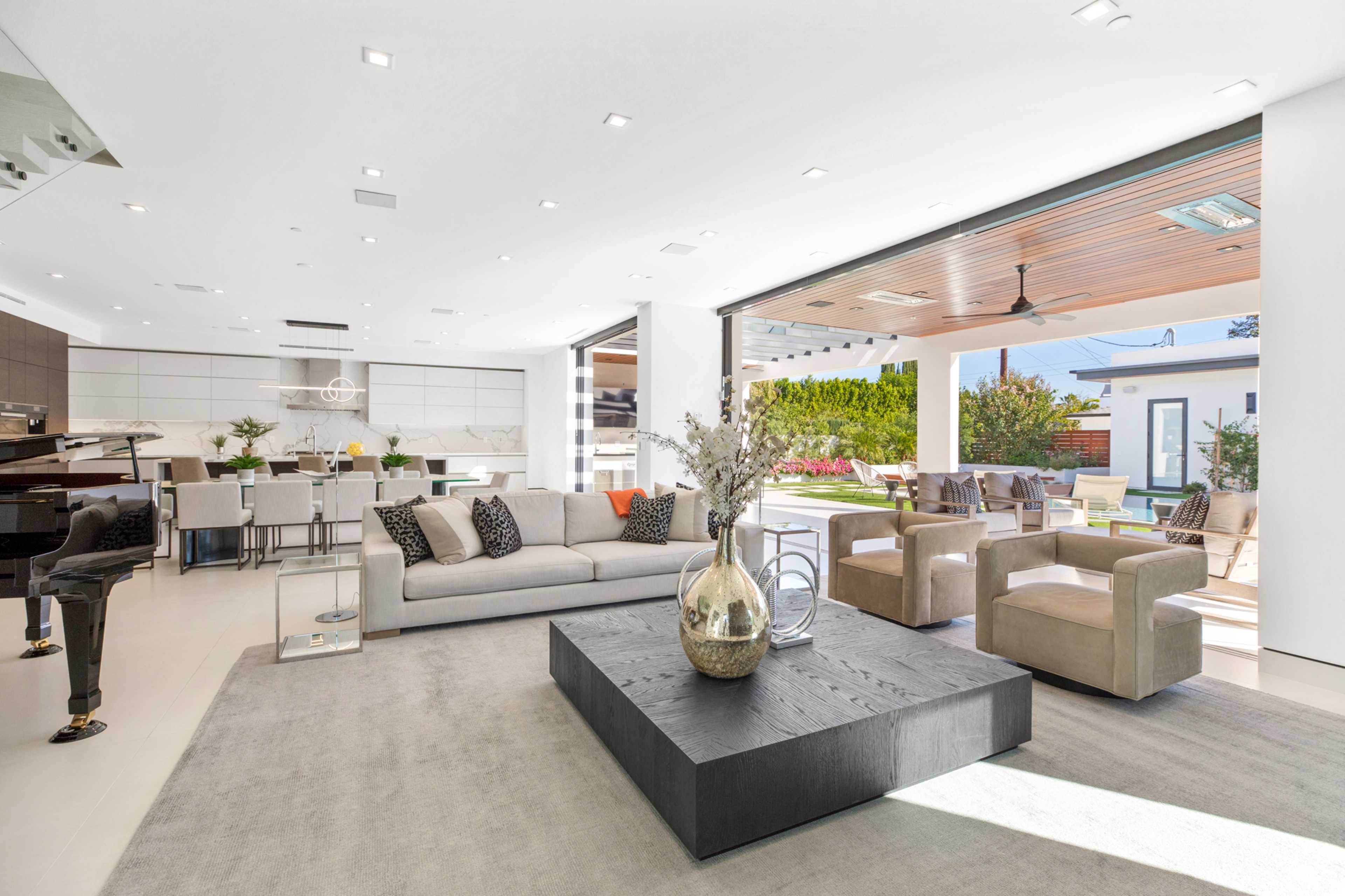 A modern living room features a large gray sofa, a coffee table, and an opening to an outdoor space with a pool and dining area.