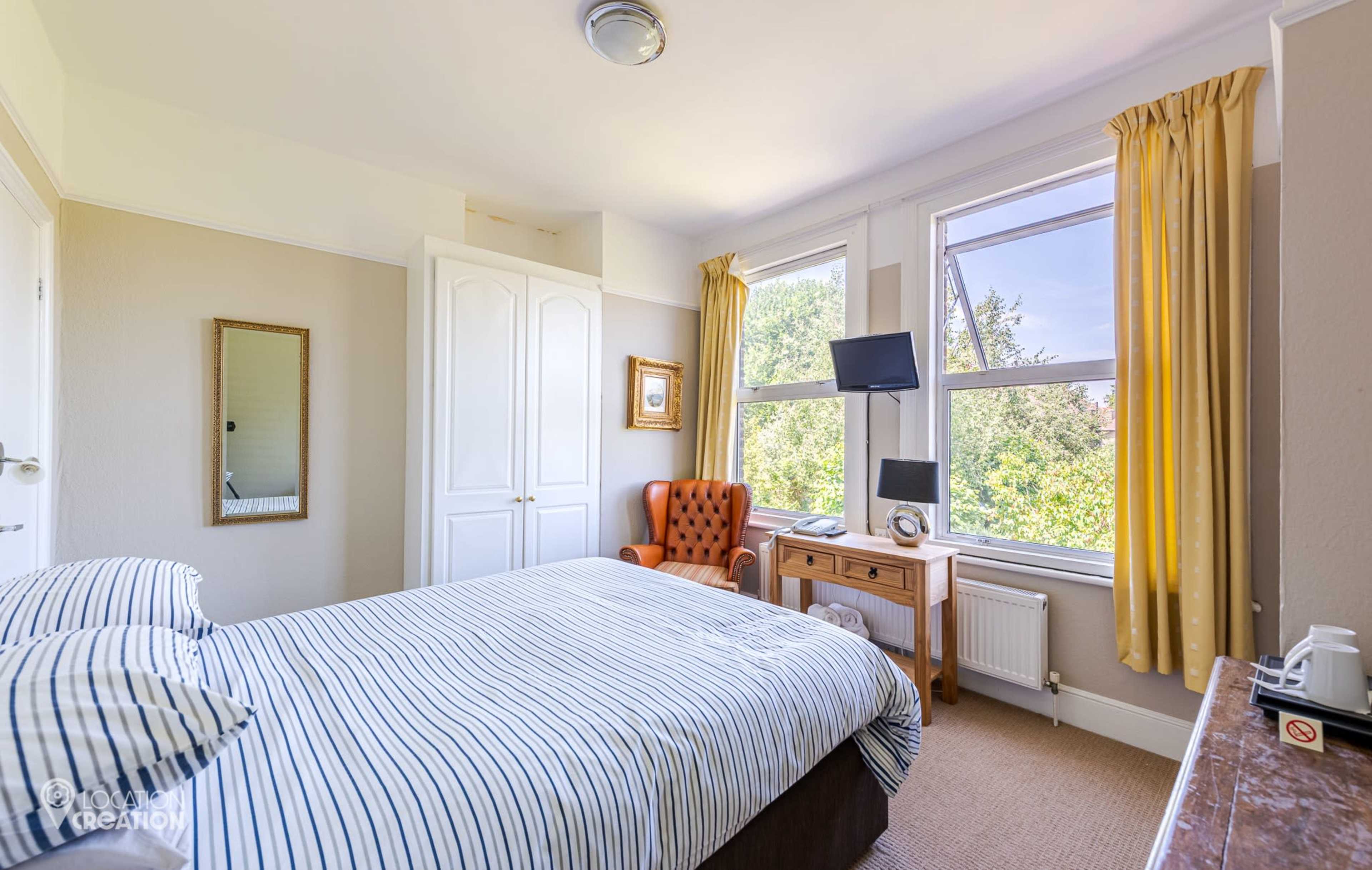 The image shows a bright bedroom with a striped bedspread, a large window with yellow curtains, and a brown armchair next to a wooden table and a flat-screen TV.