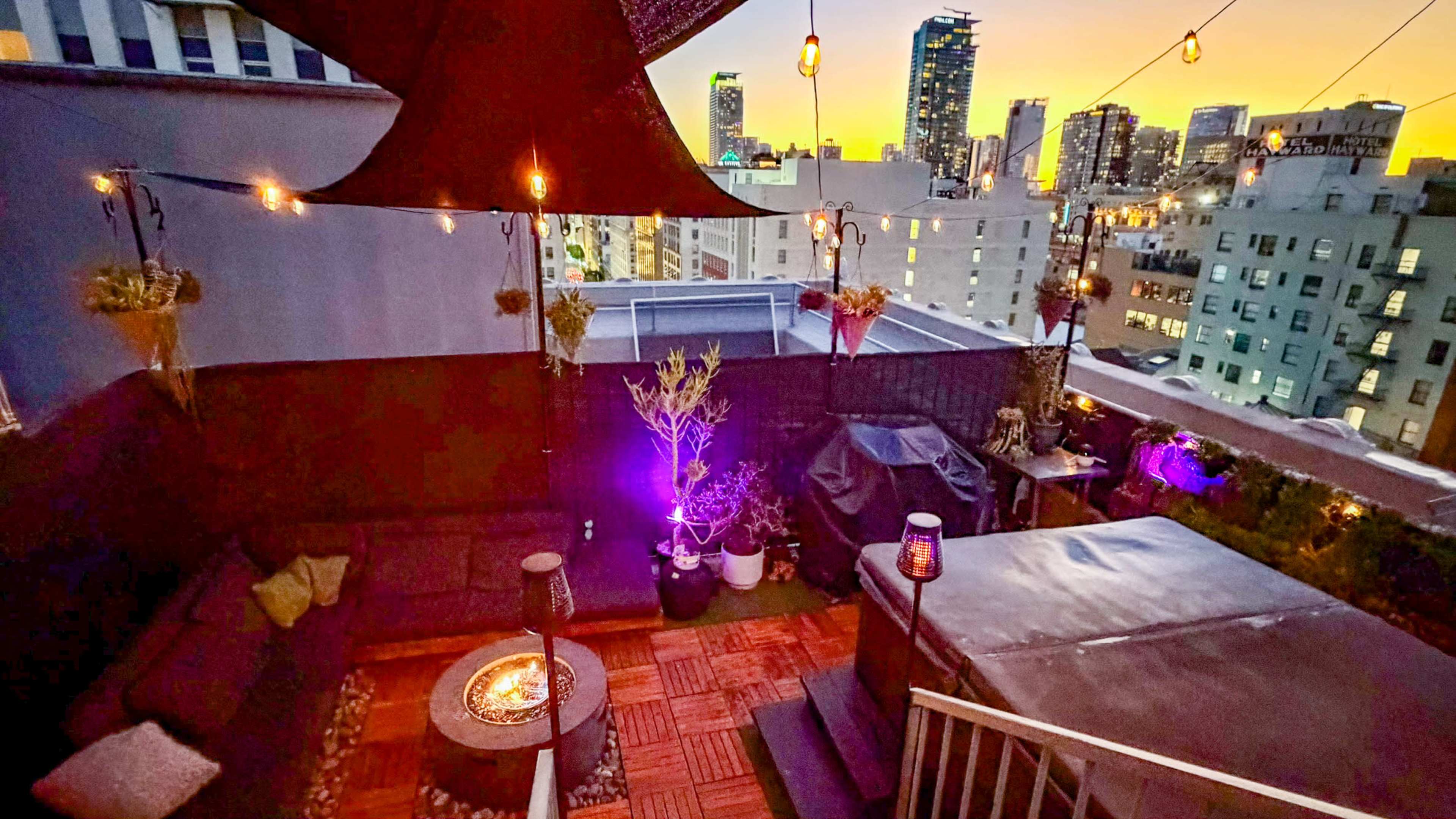 The image shows a rooftop terrace with a hot tub, seating area, and city skyline at sunset.
