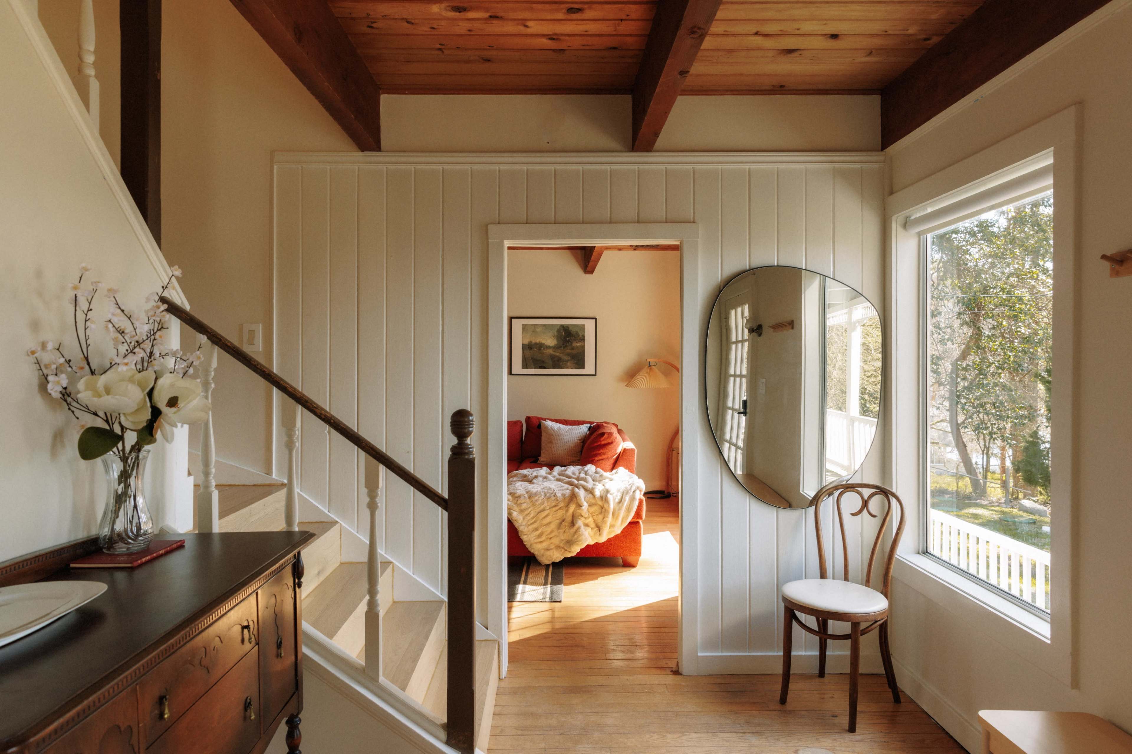A well-lit hallway leads to a cozy bedroom featuring a red couch, with a mirror and a chair positioned near a window.