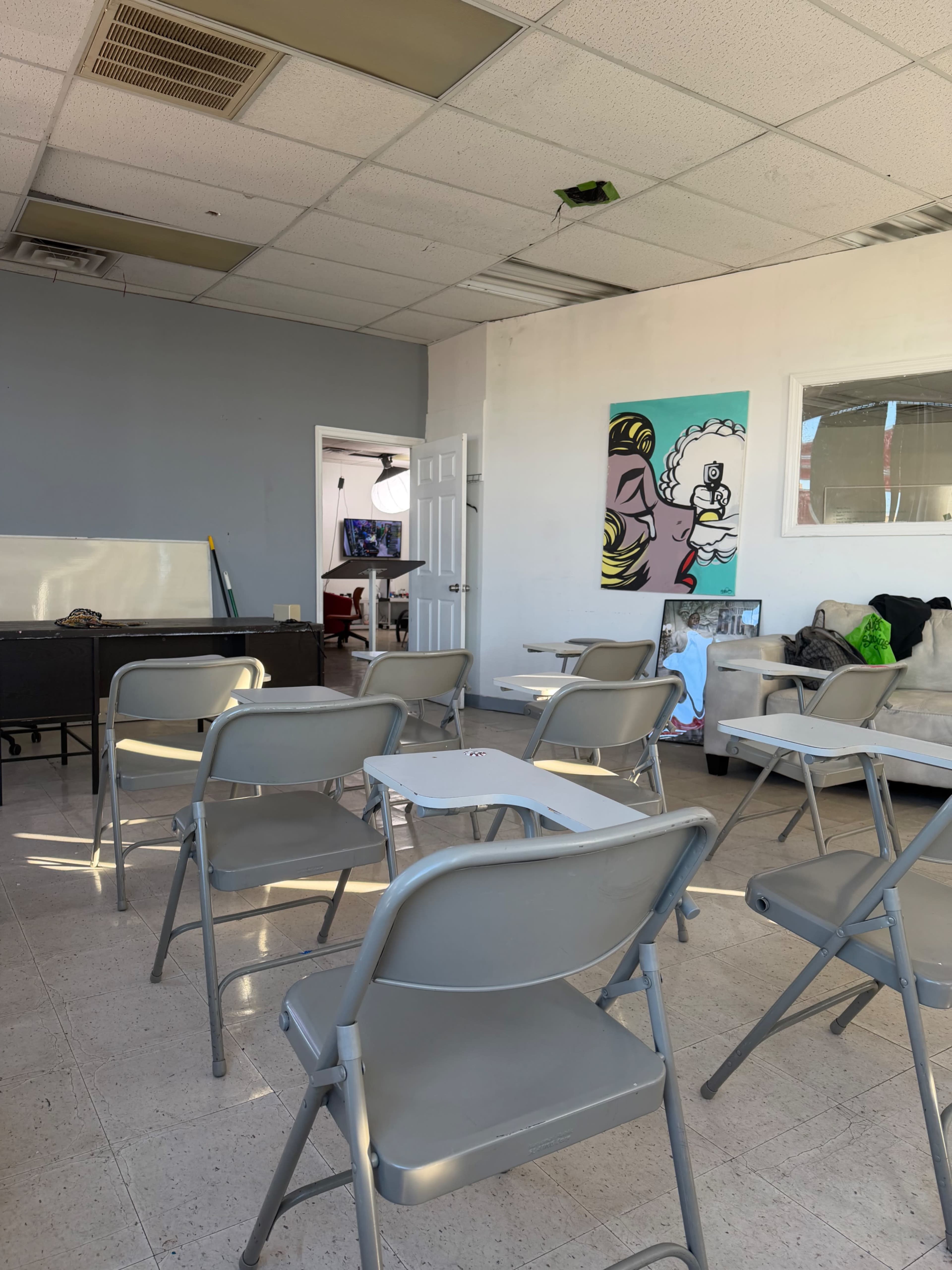The image shows a classroom setting with several gray folding chairs arranged in rows, a whiteboard, and colorful artwork on the walls.