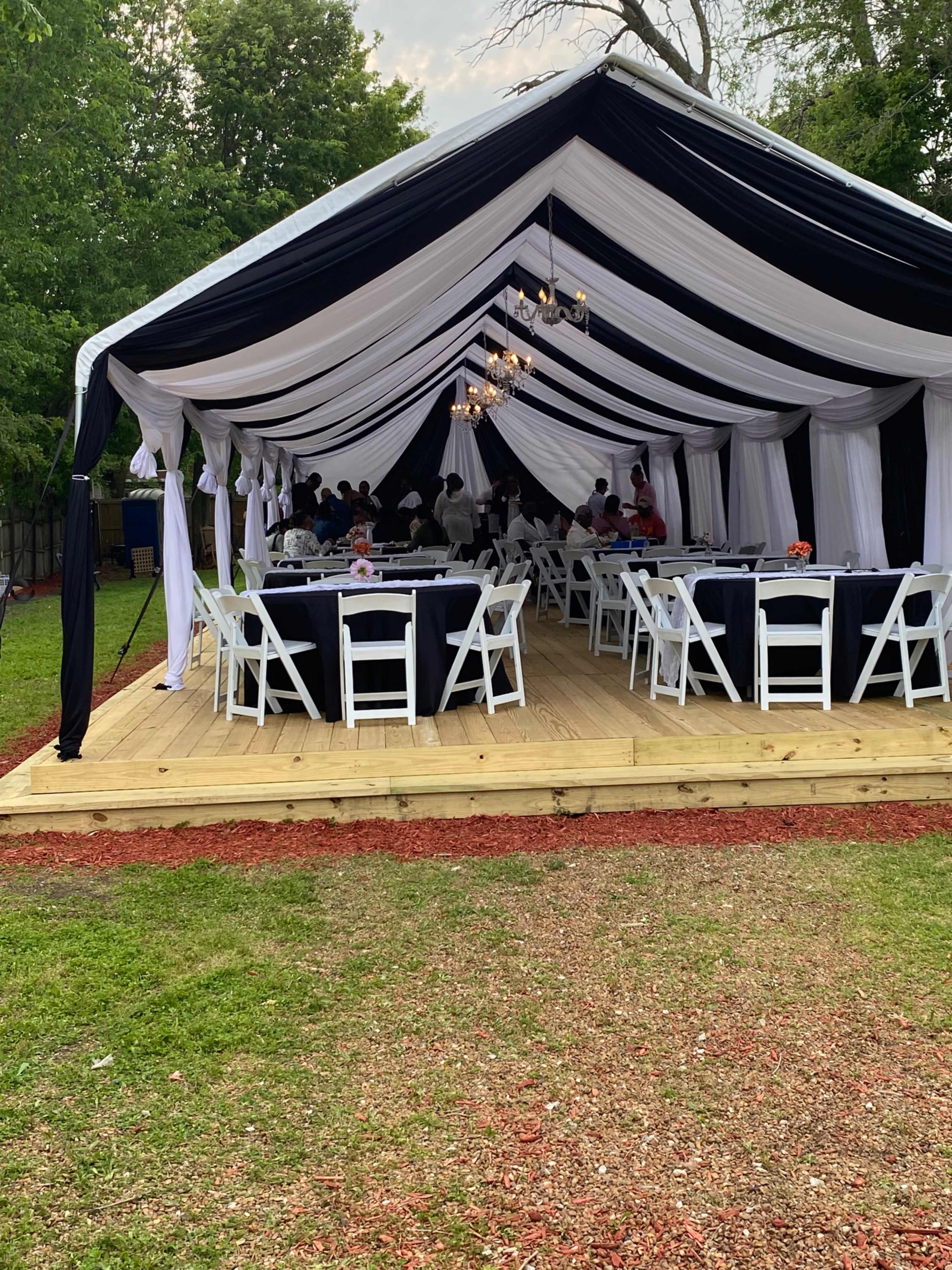 A large tent with black and white drapery is set up on a wooden platform, surrounded by green grass and red mulch, with tables and chairs arranged inside for an event.