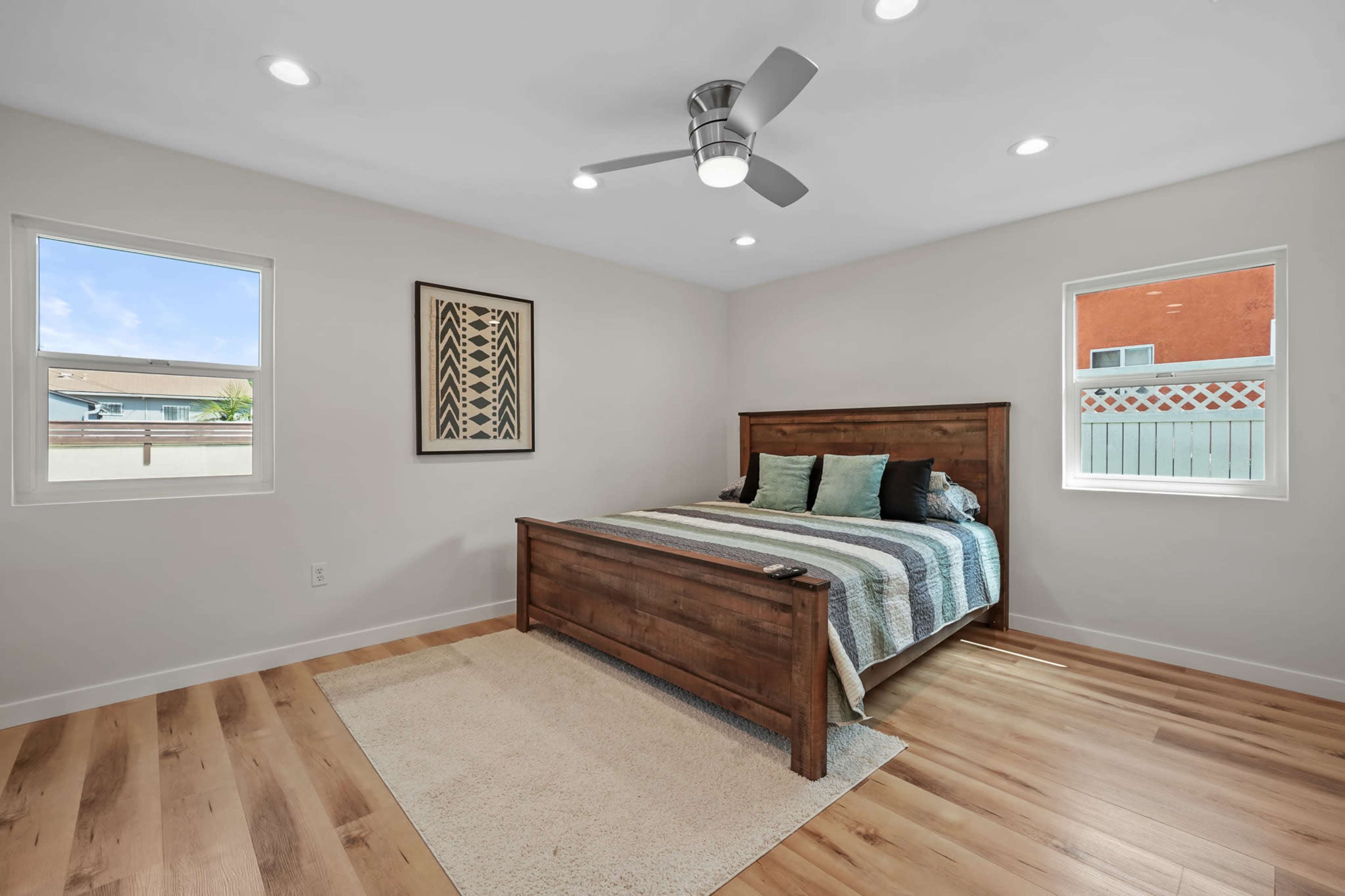 A modern bedroom featuring a wooden bed frame, patterned bedding, two windows allowing natural light, and a ceiling fan.
