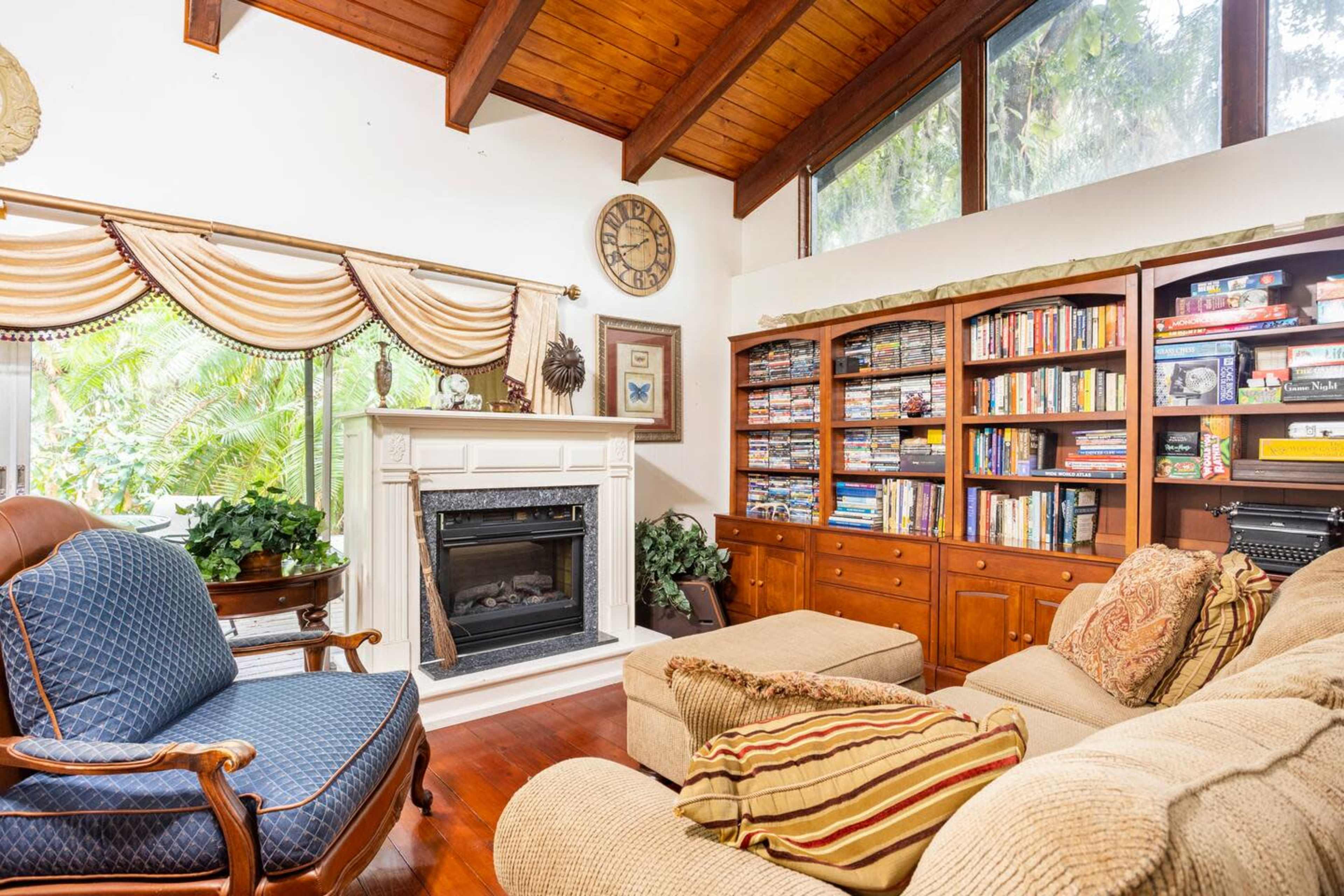 The image shows a cozy living room featuring a fireplace, a large bookshelf filled with books and games, and comfortable seating arranged around a coffee table.