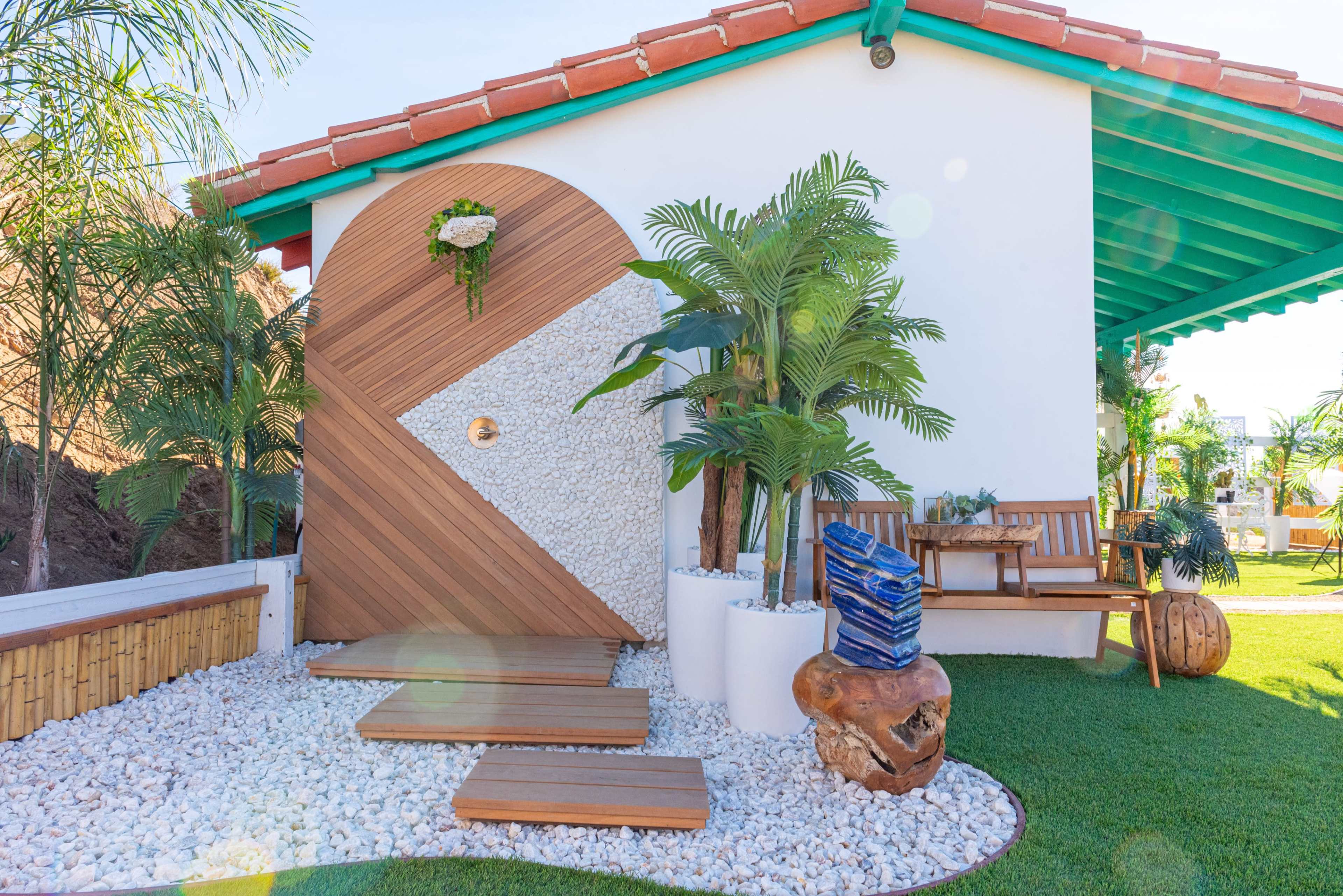 The image shows a modern outdoor space featuring a textured wooden wall, a circular window, decorative stones, and lush greenery, with wooden seating arranged nearby.