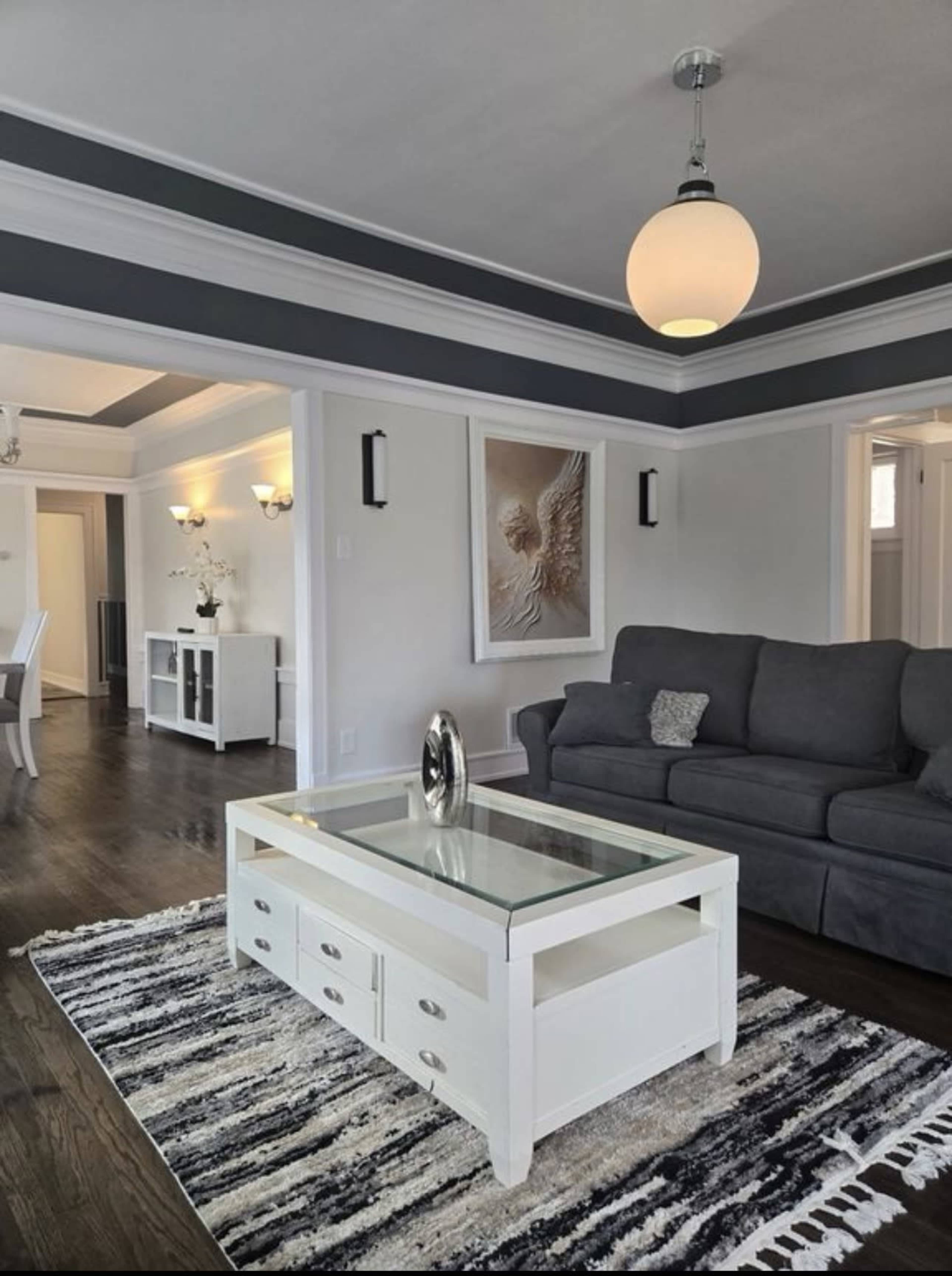 The image shows a modern living room featuring a gray sofa, a glass-top coffee table with drawers, and a patterned rug under a ceiling light fixture.