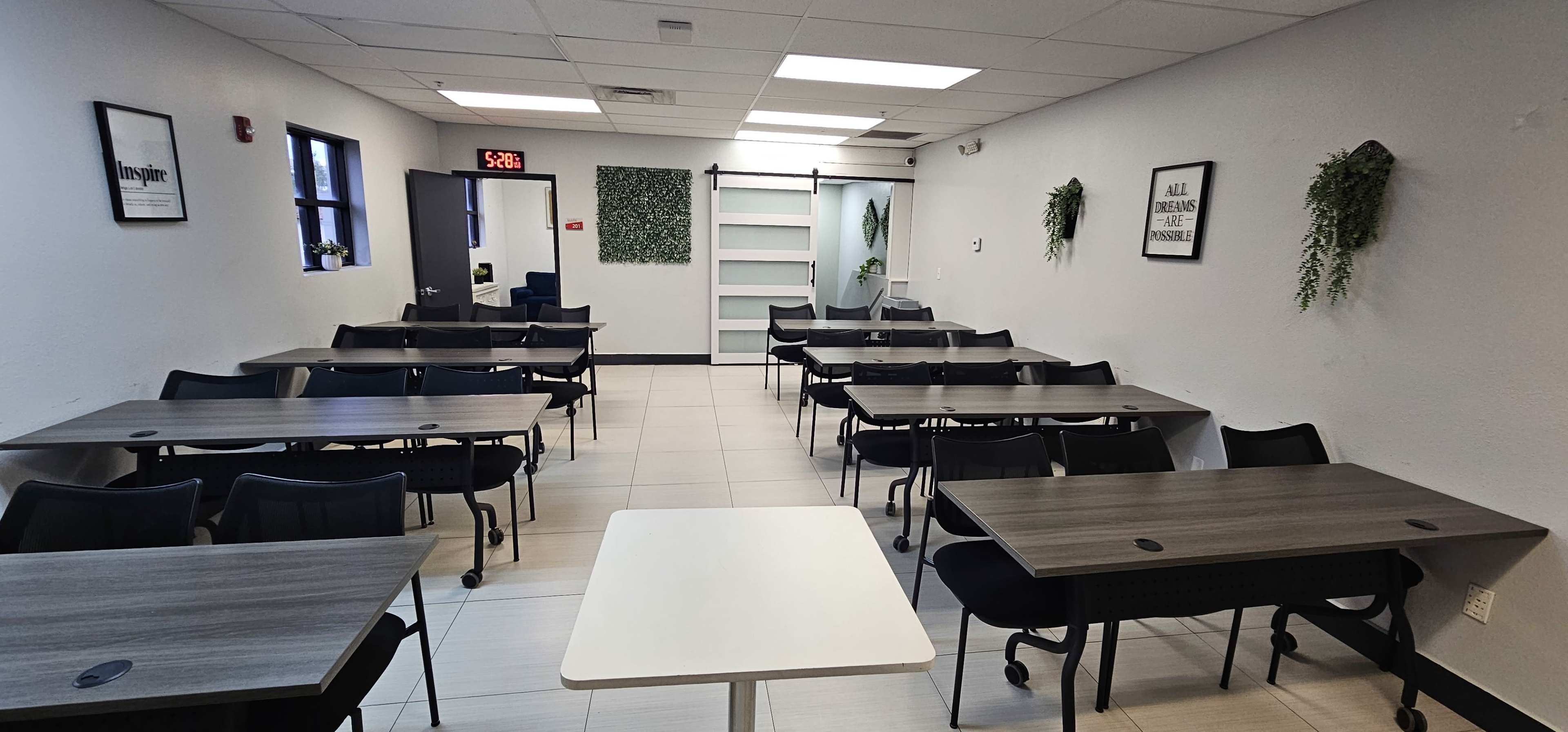 The image shows a classroom setting with several rectangular tables arranged in rows, surrounded by chairs, and well-lit by overhead lights.