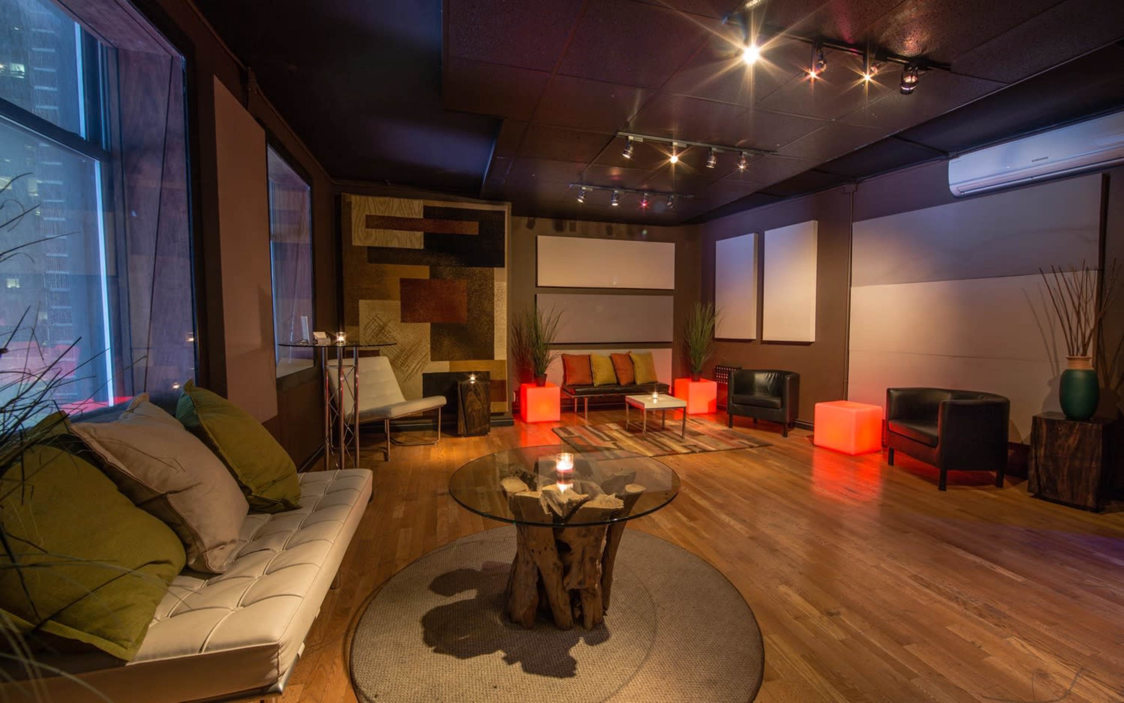 The image shows a modern, well-lit lounge with a mix of seating arrangements, including a white sofa and black chairs, centered around a glass coffee table on a circular rug.