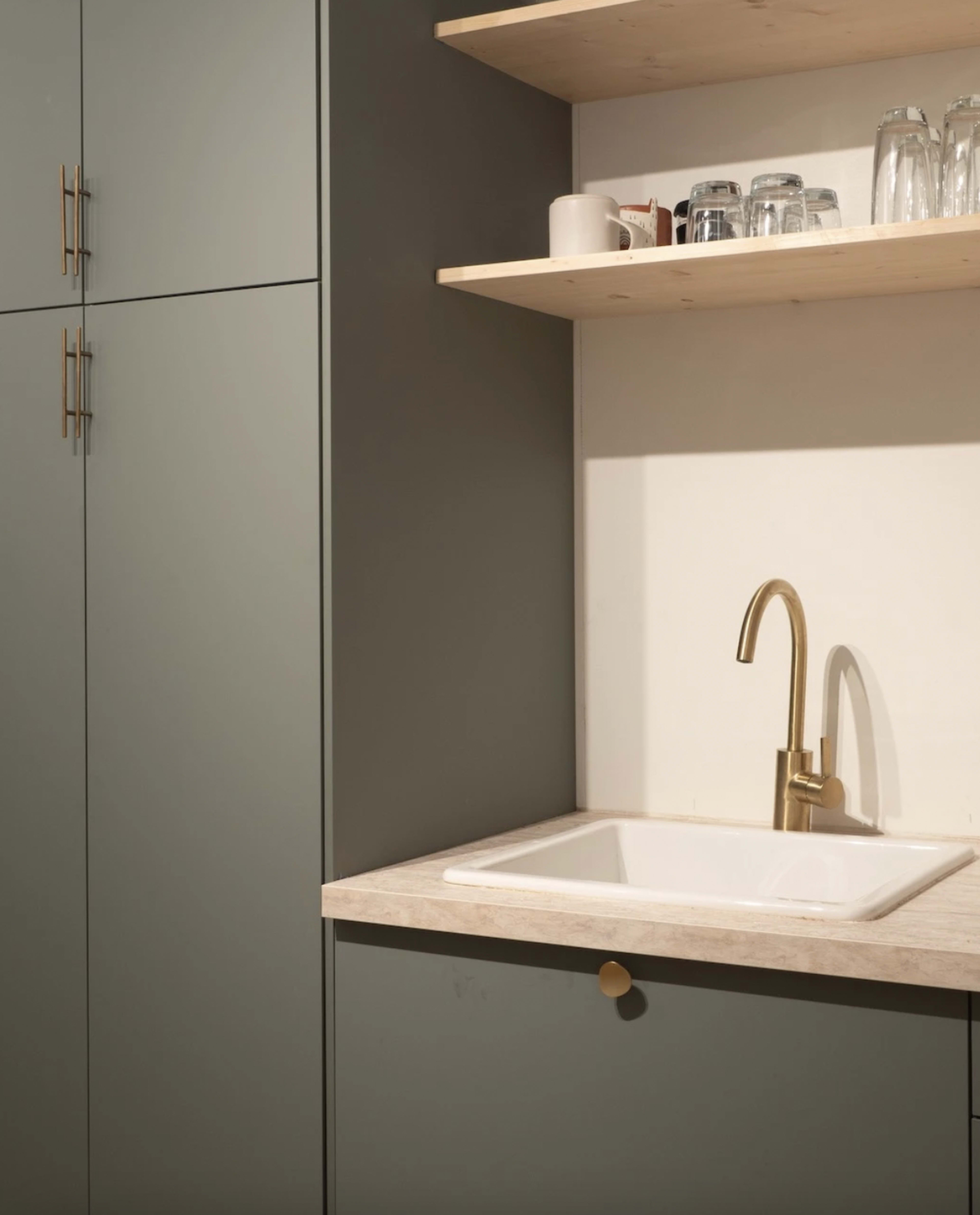 A kitchen corner with a sink, a brass faucet, and upper shelves holding glassware, all set against a muted green cabinetry backdrop.