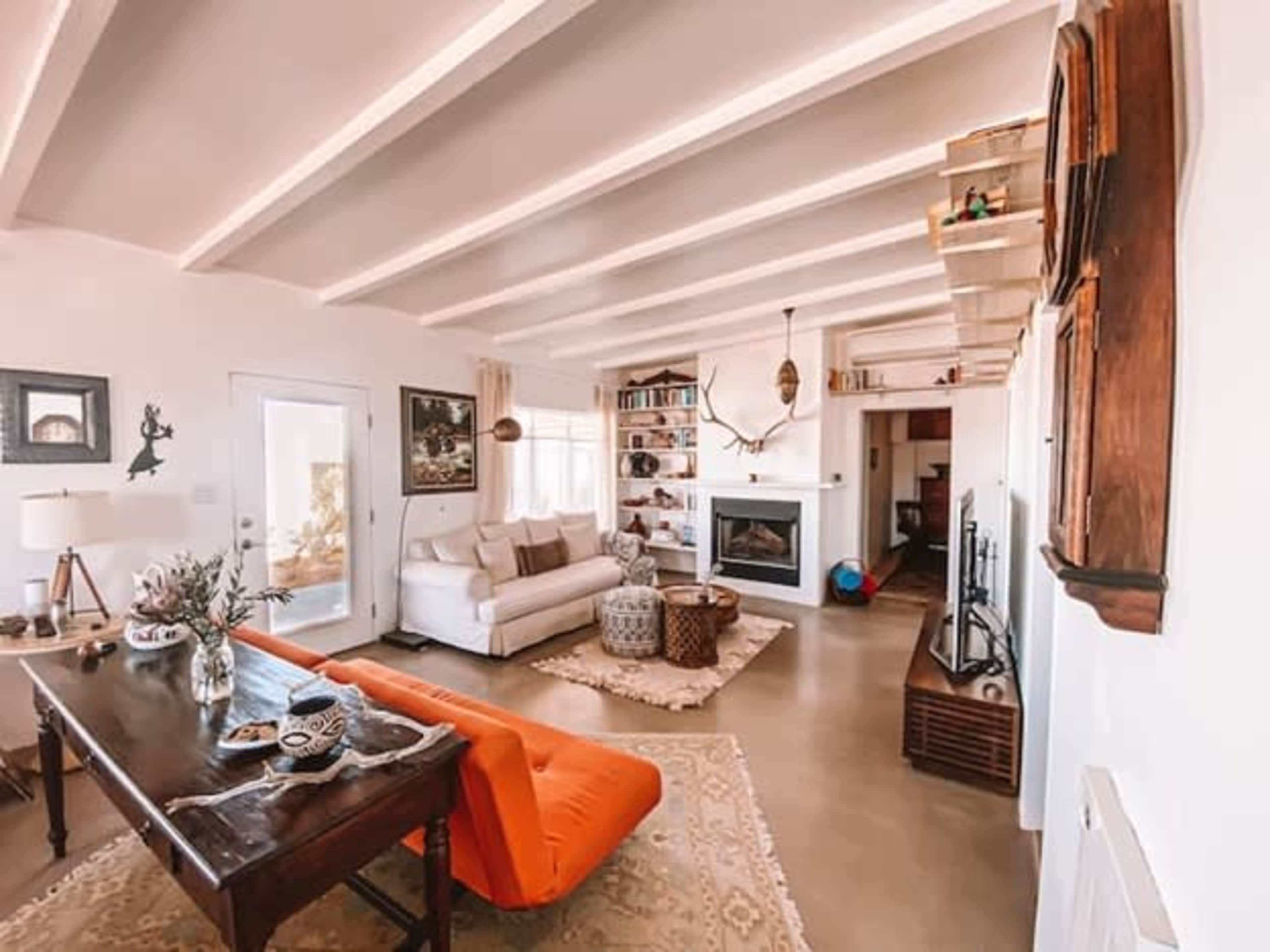 The living room features a white couch, an orange lounge chair, a wooden dining table, and a fireplace, with shelves and decorations along the walls.