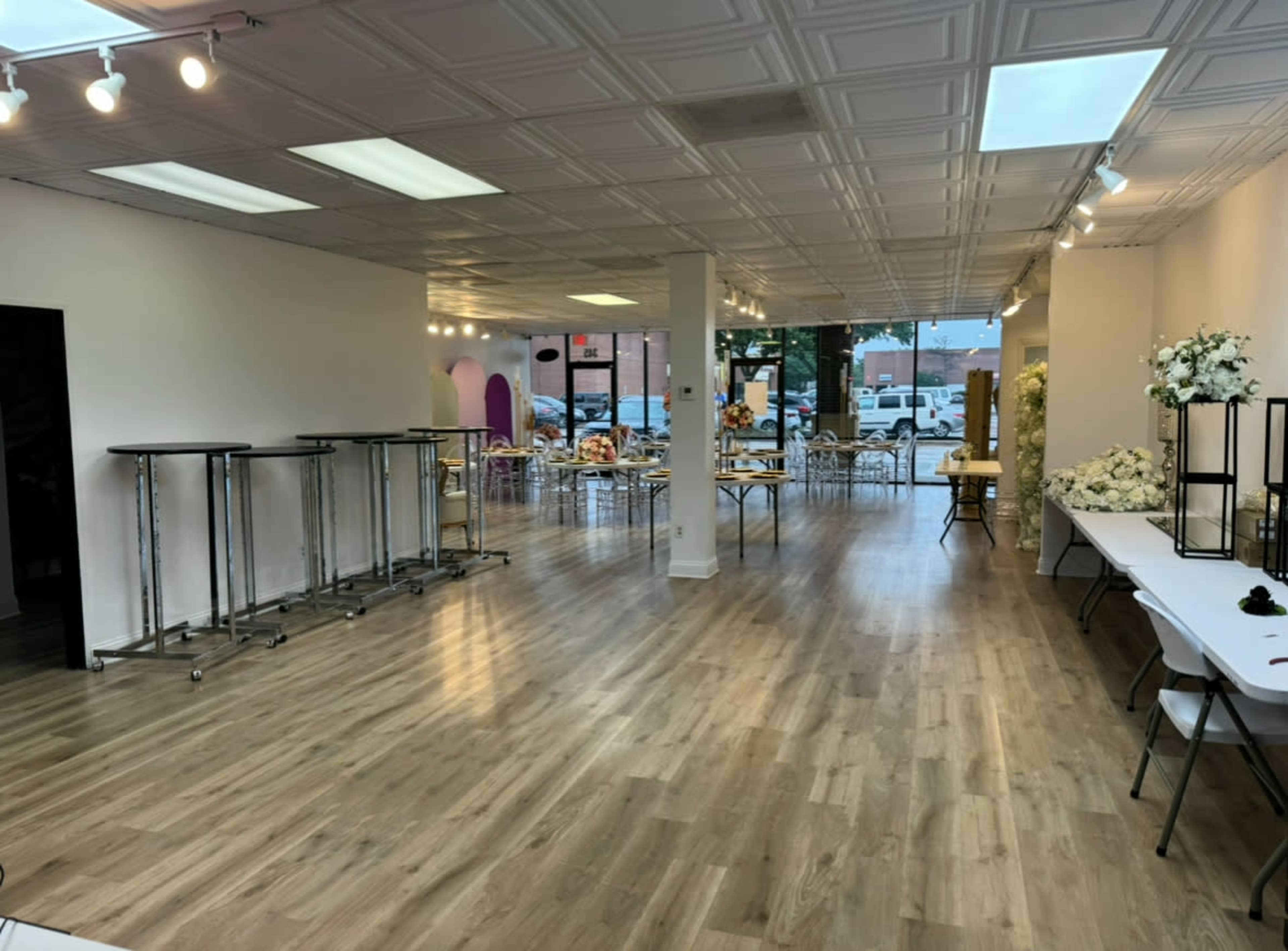 The image shows a spacious, well-lit interior of a venue with wooden flooring, folding tables, and empty standing tables arranged throughout.