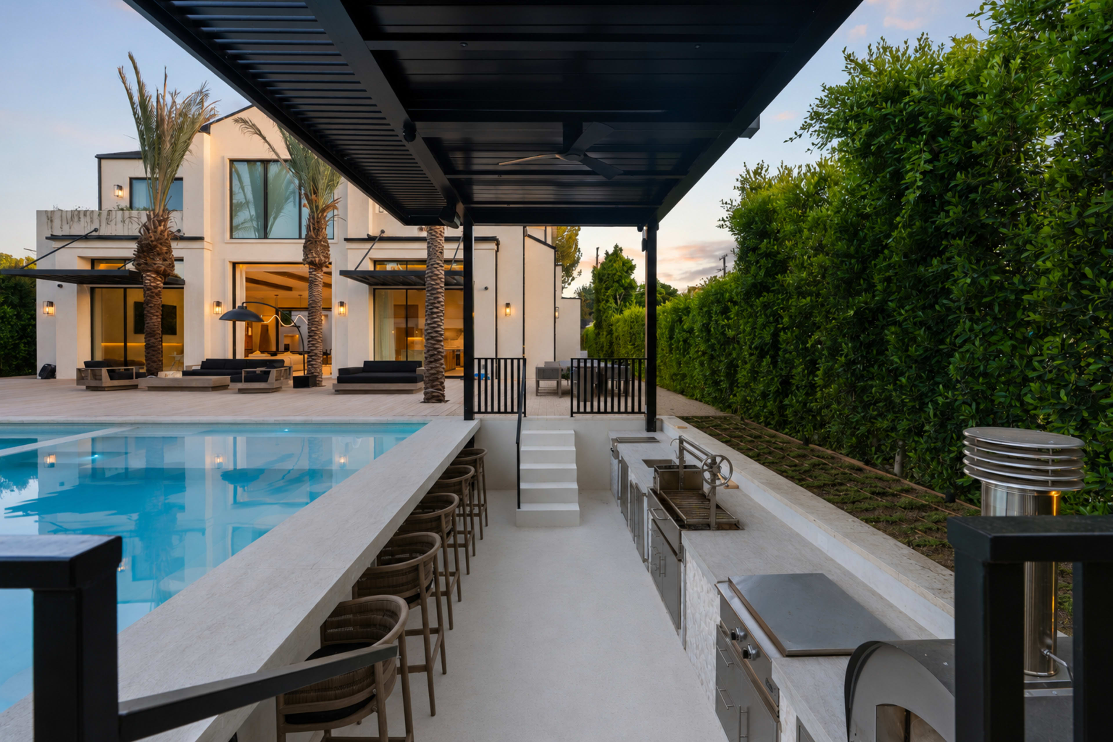 The image shows a modern outdoor kitchen and pool area adjacent to a contemporary two-story house, surrounded by tall palm trees and lush greenery.