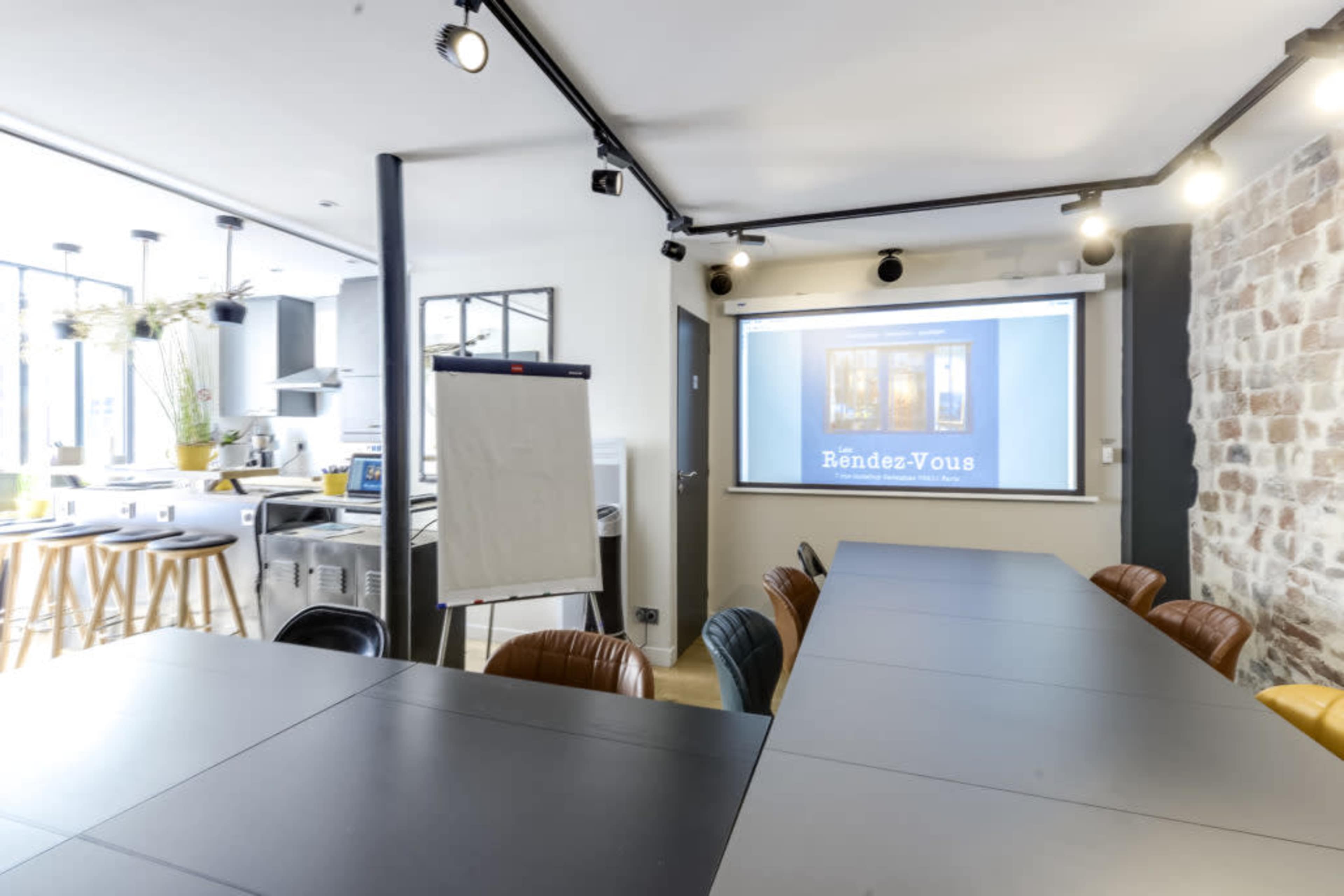 A modern conference room features a long table, a projector screen displaying "Rendez-Vous," and a kitchen area visible through large windows.