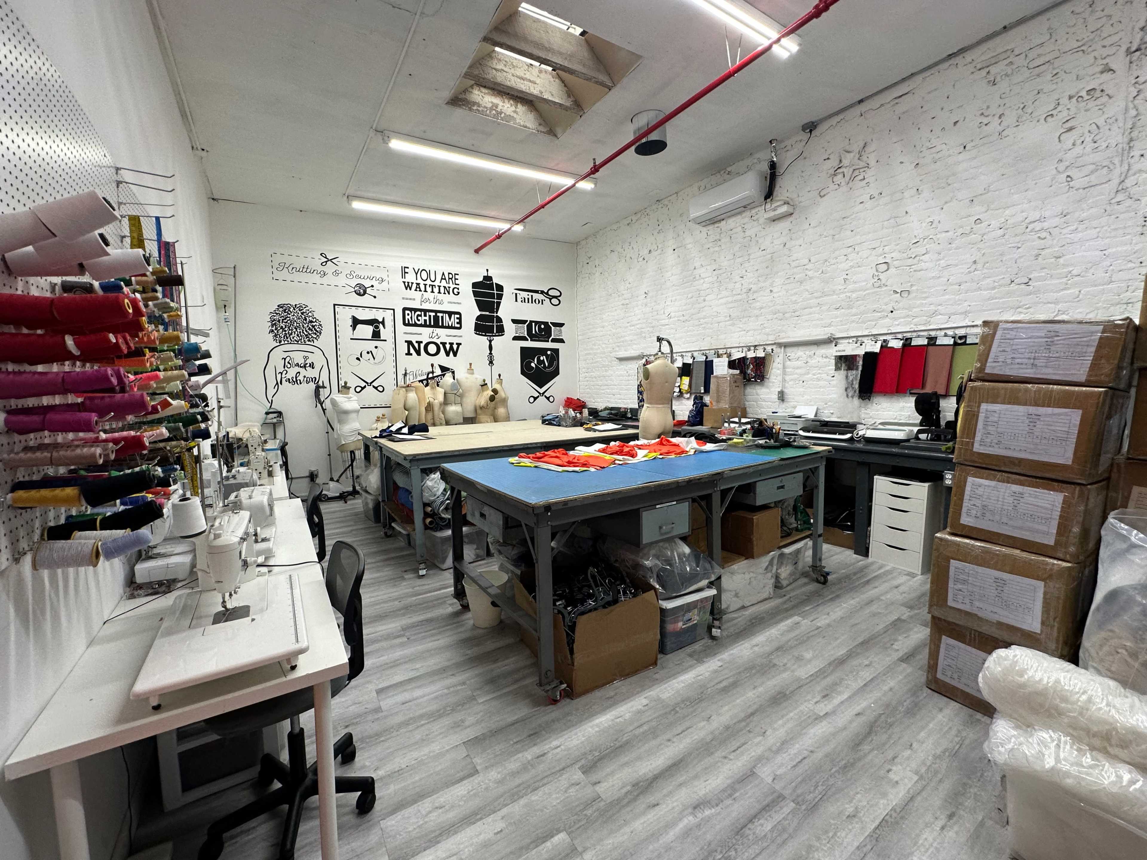 A workshop space features sewing machines, tables with fabric, and stacks of boxes against the walls adorned with various designs and text.
