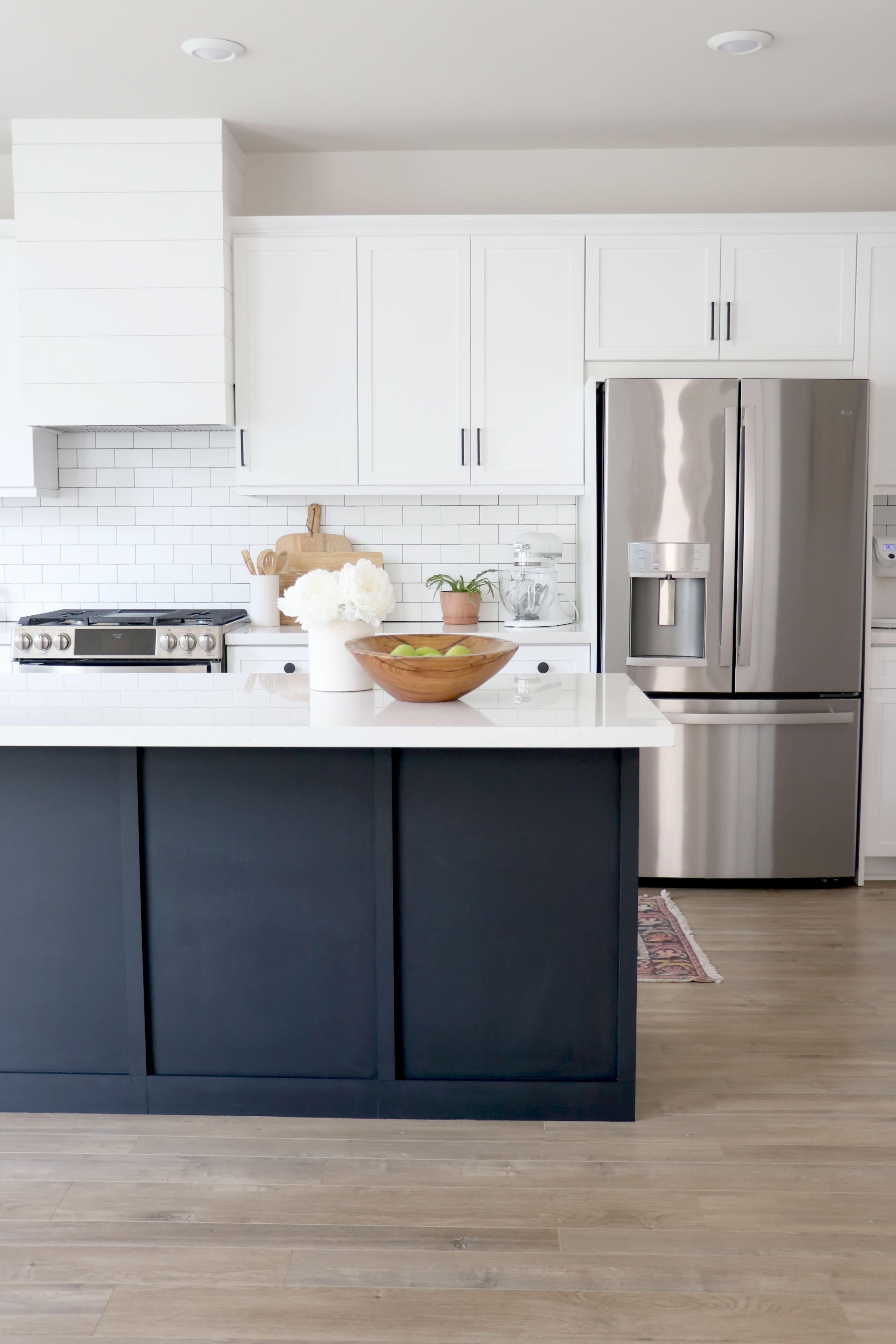 A modern kitchen with white cabinets, a stainless steel refrigerator, and a dark blue island featuring a wooden bowl of green apples.