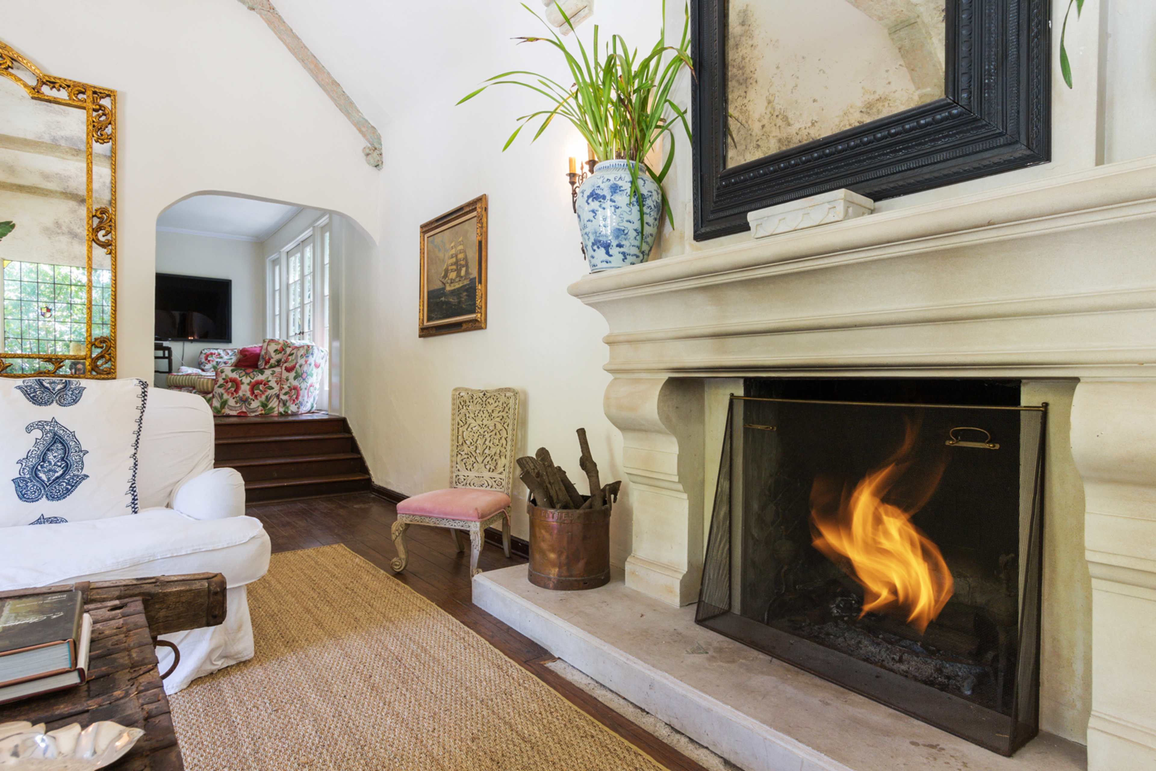 A cozy living room features a stone fireplace with a visible flame, surrounded by a white sofa, decorative plants, and artwork on the walls.