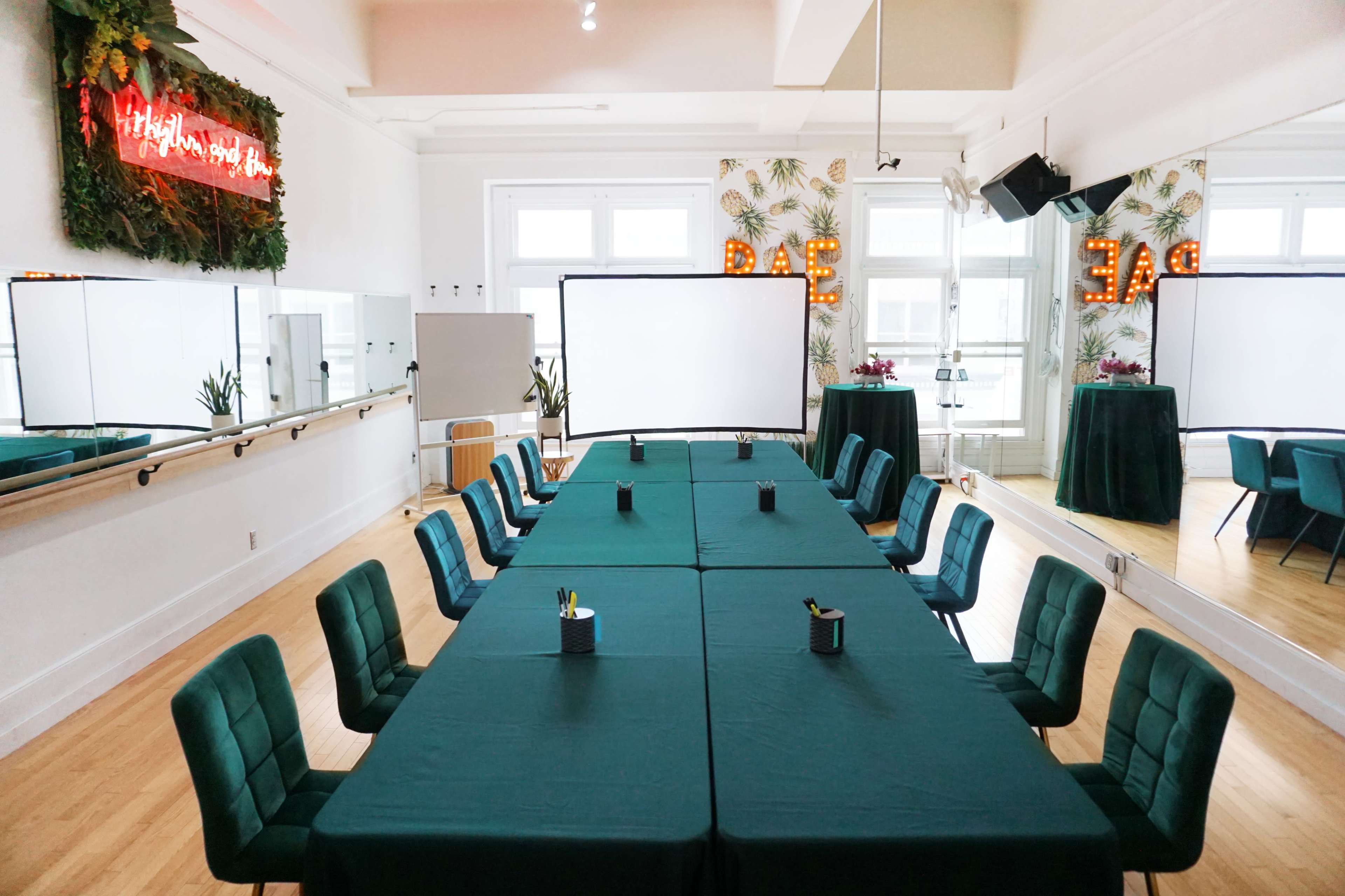 A meeting room features two long tables arranged in an L-shape, covered with green tablecloths, surrounded by teal chairs, with a decorative backdrop and a blank screen at one end.