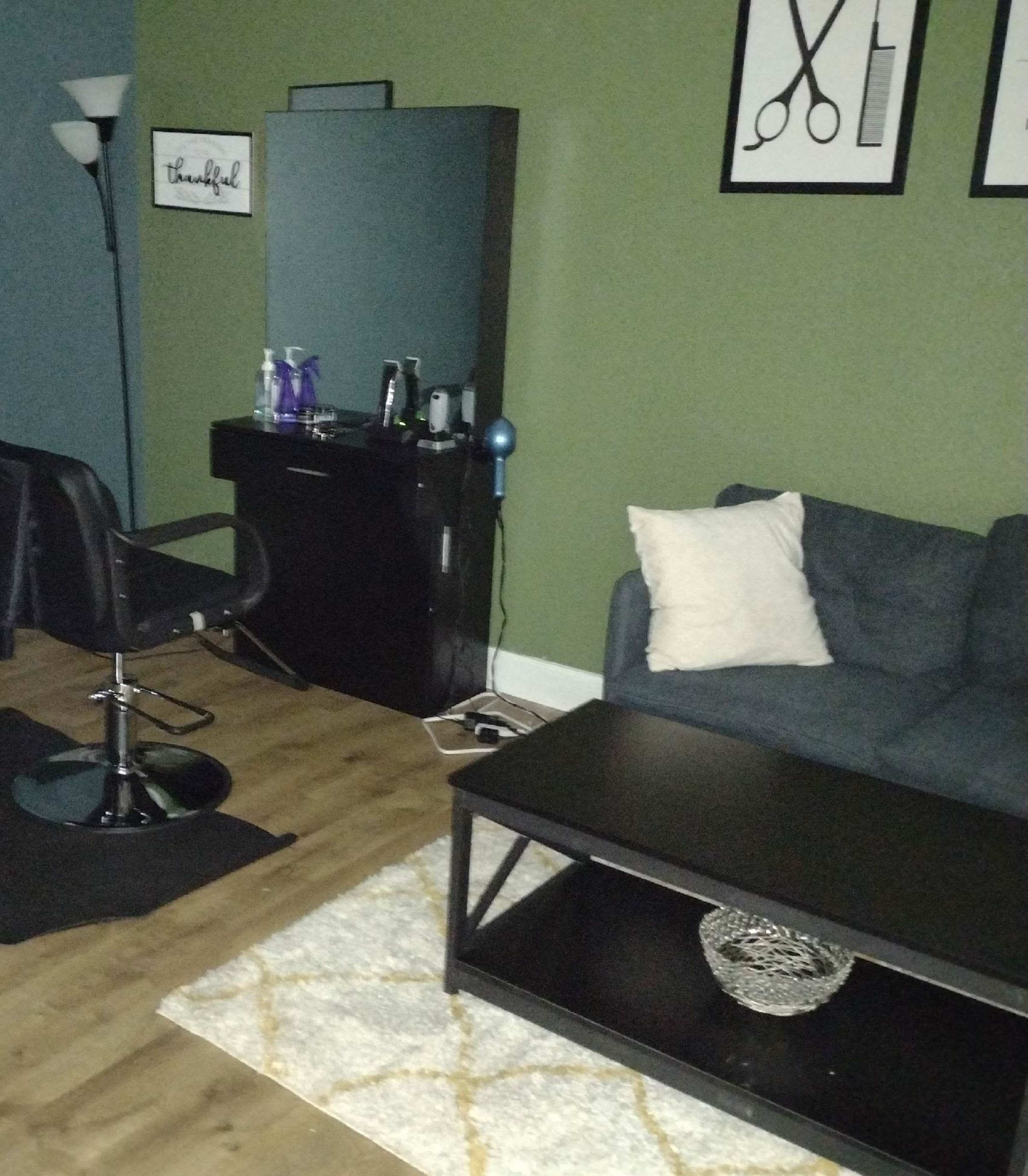 A dimly lit room with a black salon chair, a dark cabinet, a blue lamp, and a gray couch on a light-colored rug.