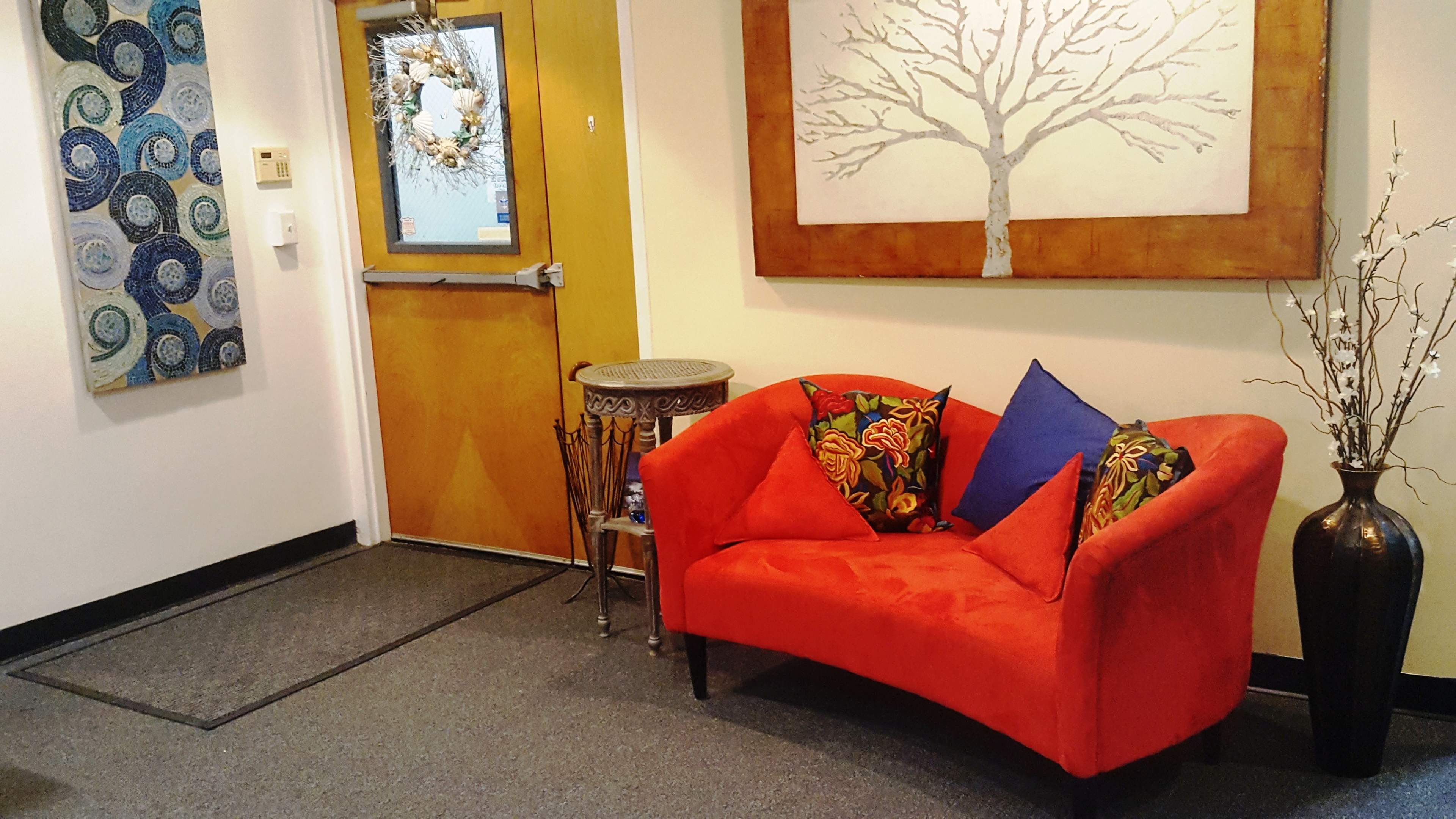 A small entryway featuring a red couch with patterned cushions, a wooden door with a wreath, and a decorative vase next to a wall art piece depicting a tree.