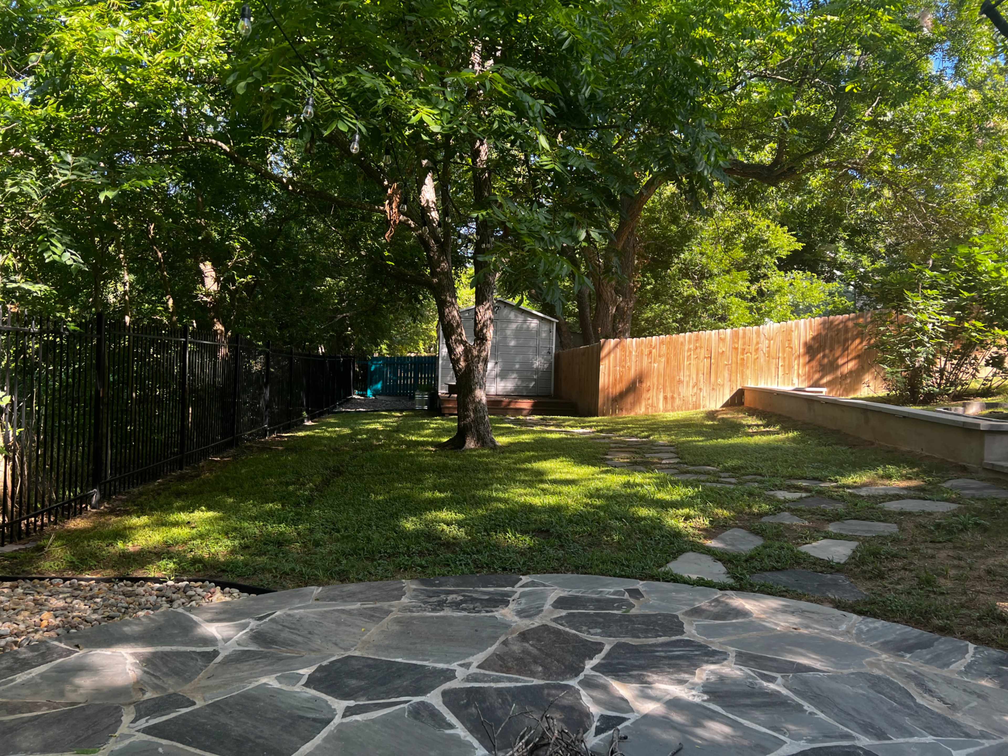 A backyard features a stone patio, grassy areas, trees, and a wooden fence in the background.