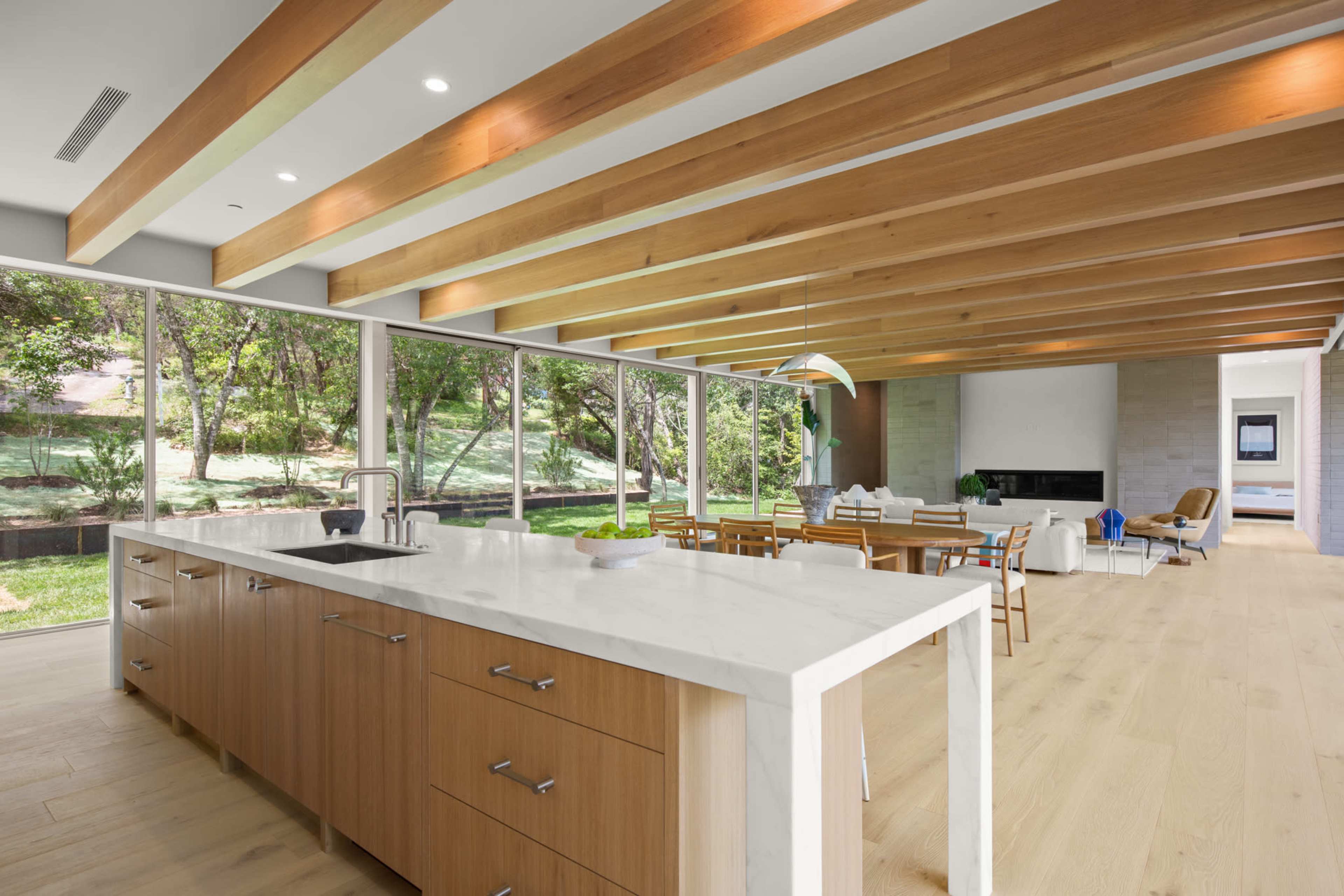 A modern kitchen with a large island and wooden beams overlooks a spacious living area filled with natural light and greenery outside.