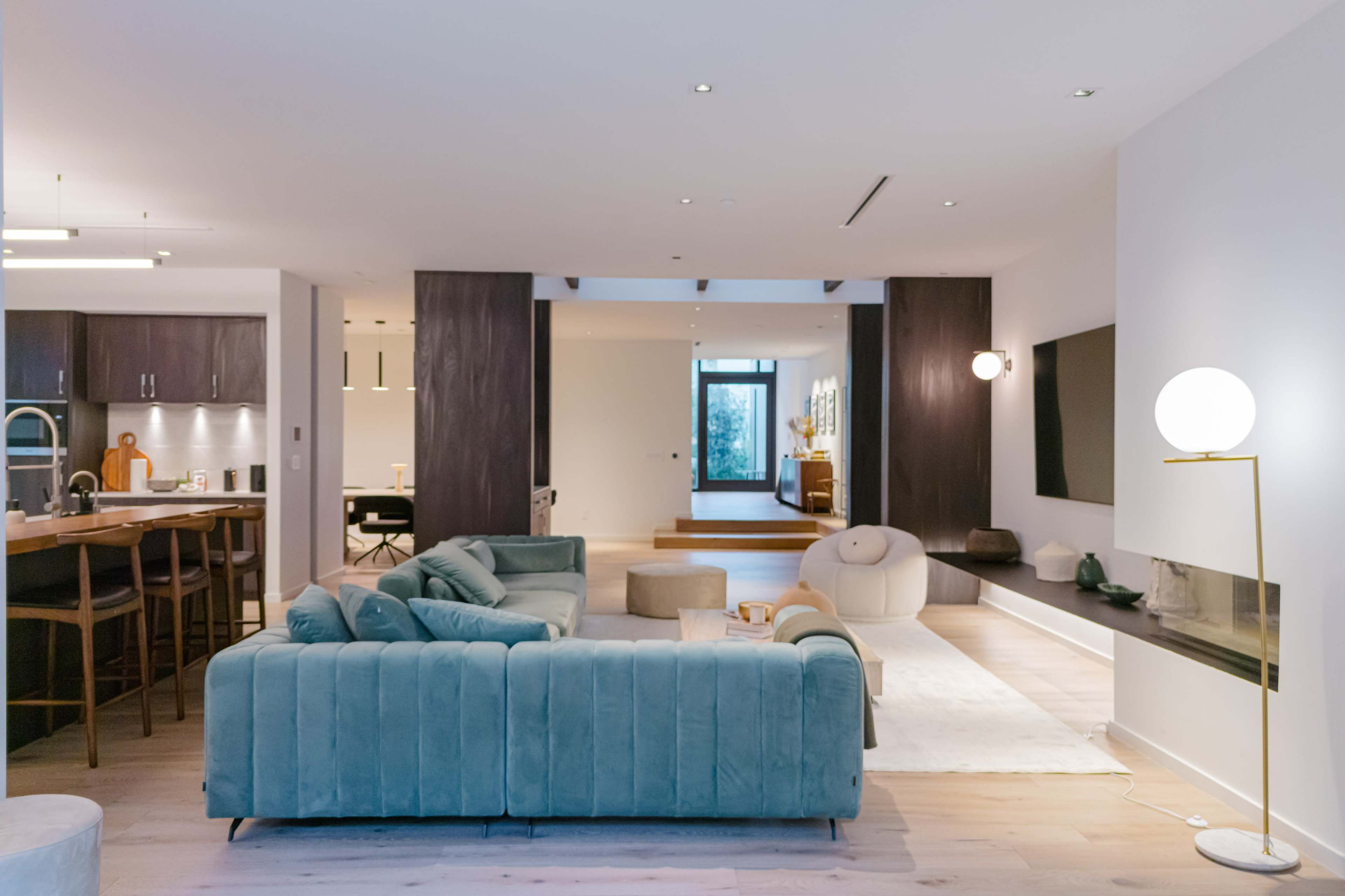 A modern, open-concept living space featuring a green sofa, a kitchen area with bar stools, and a light-colored rug on a wooden floor.