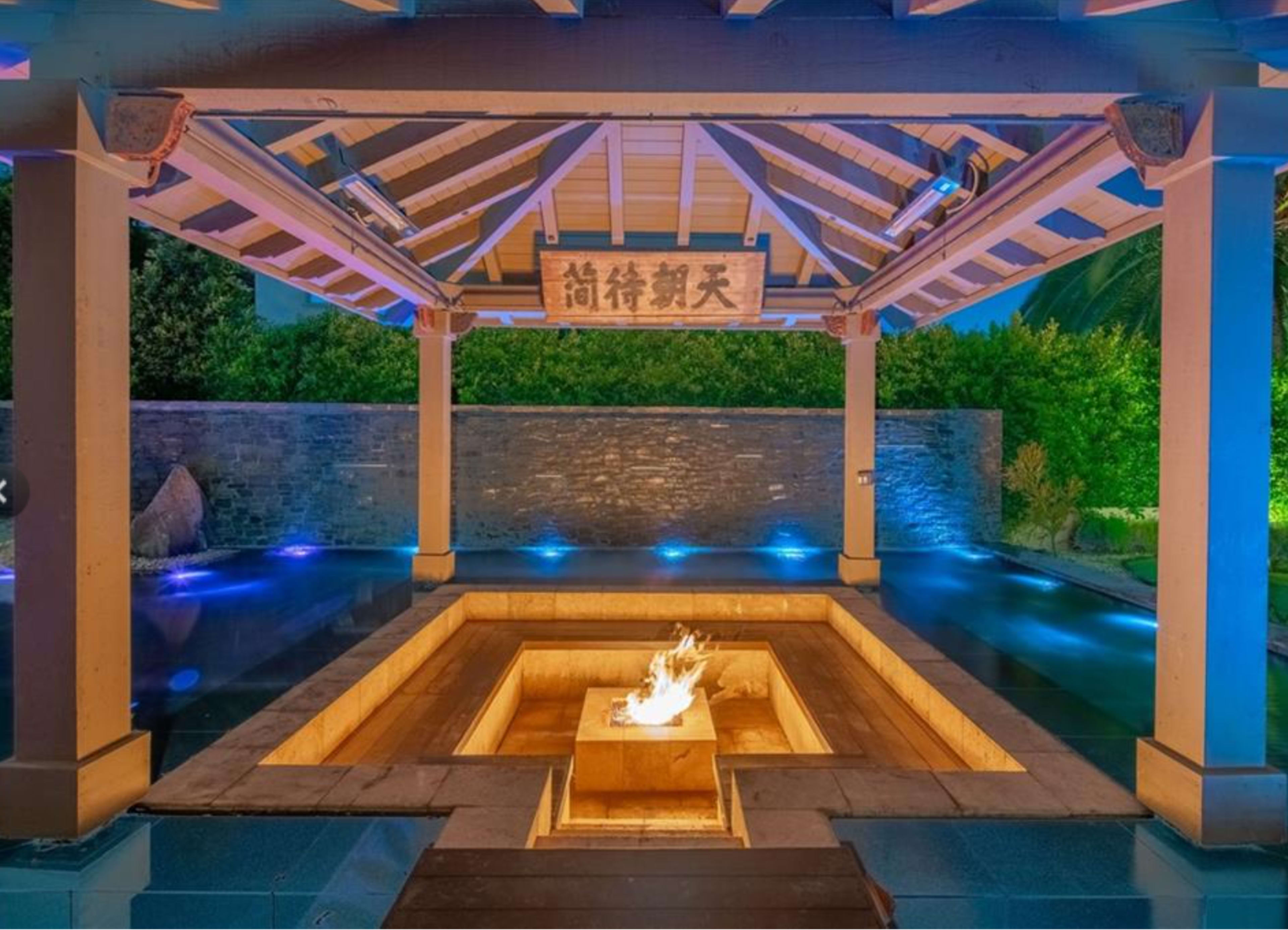 The image shows a covered outdoor space featuring a square fire pit at the center, surrounded by water and illuminated by blue lights, with a decorative sign overhead.