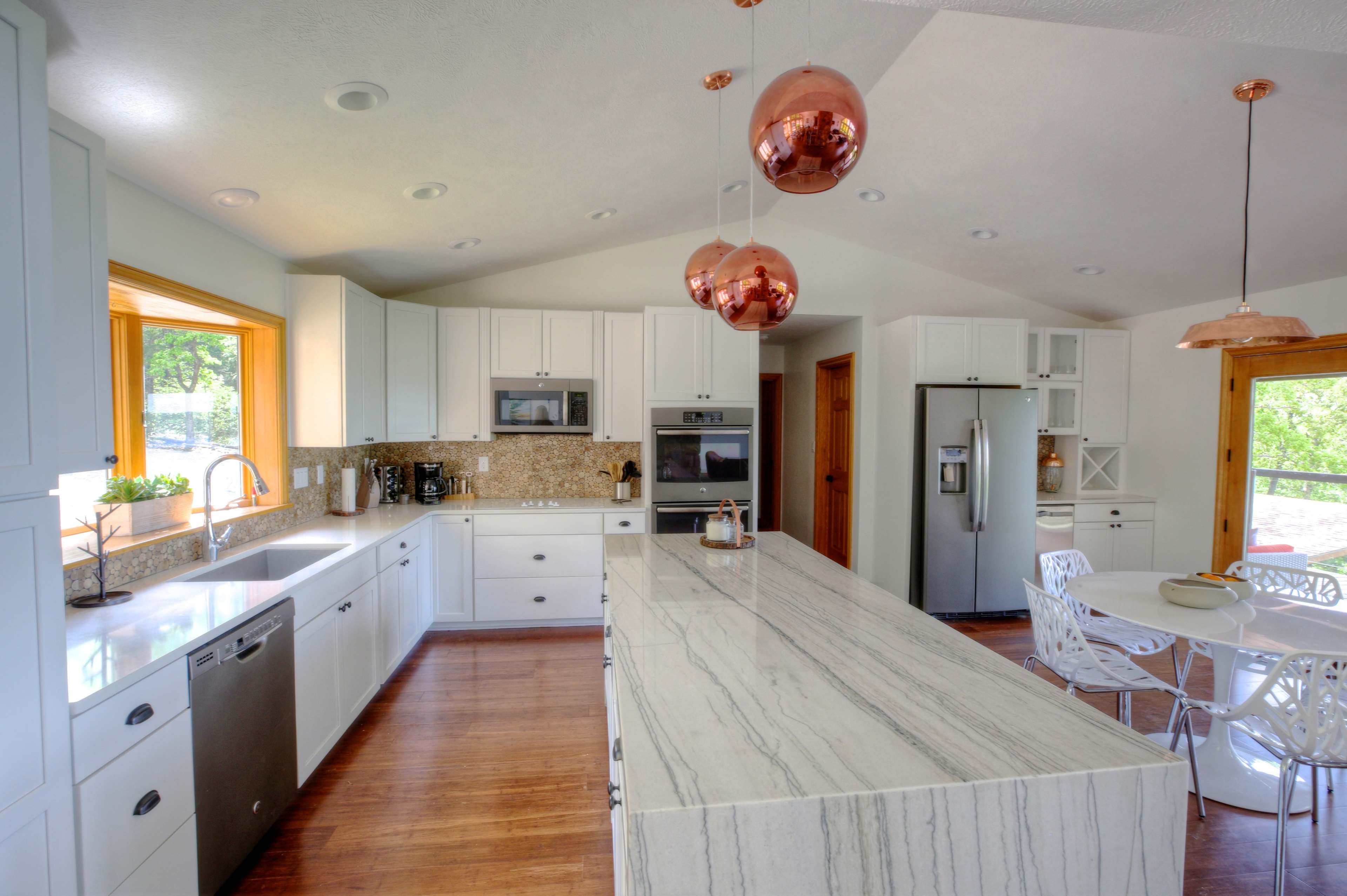 The image shows a modern kitchen with white cabinets, a large marble island, and copper pendant lights, leading to a dining area with a round table and chairs.
