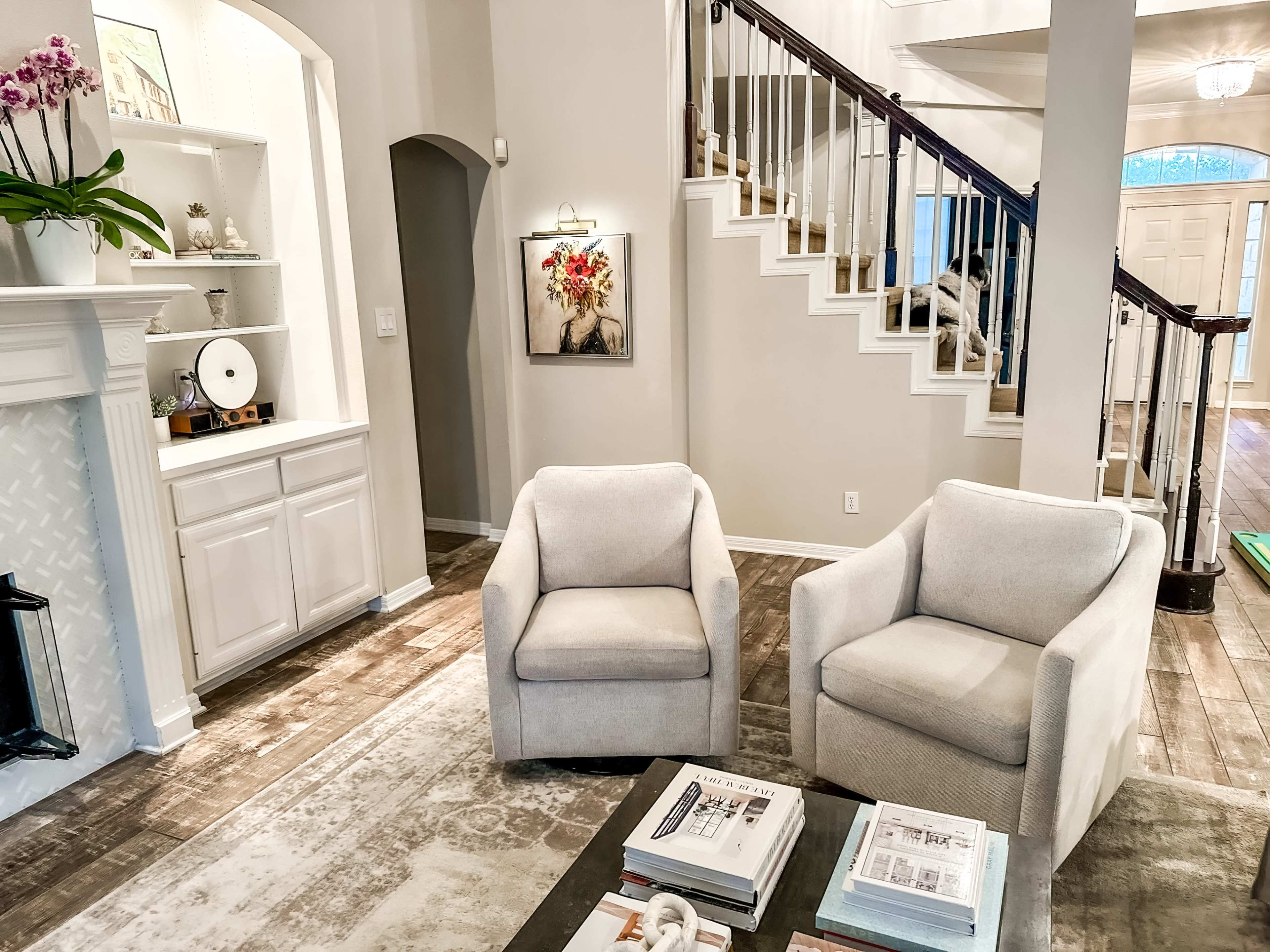 The image shows a living room with two light-colored armchairs positioned around a dark coffee table, featuring a staircase in the background and decorative elements on a nearby shelf.