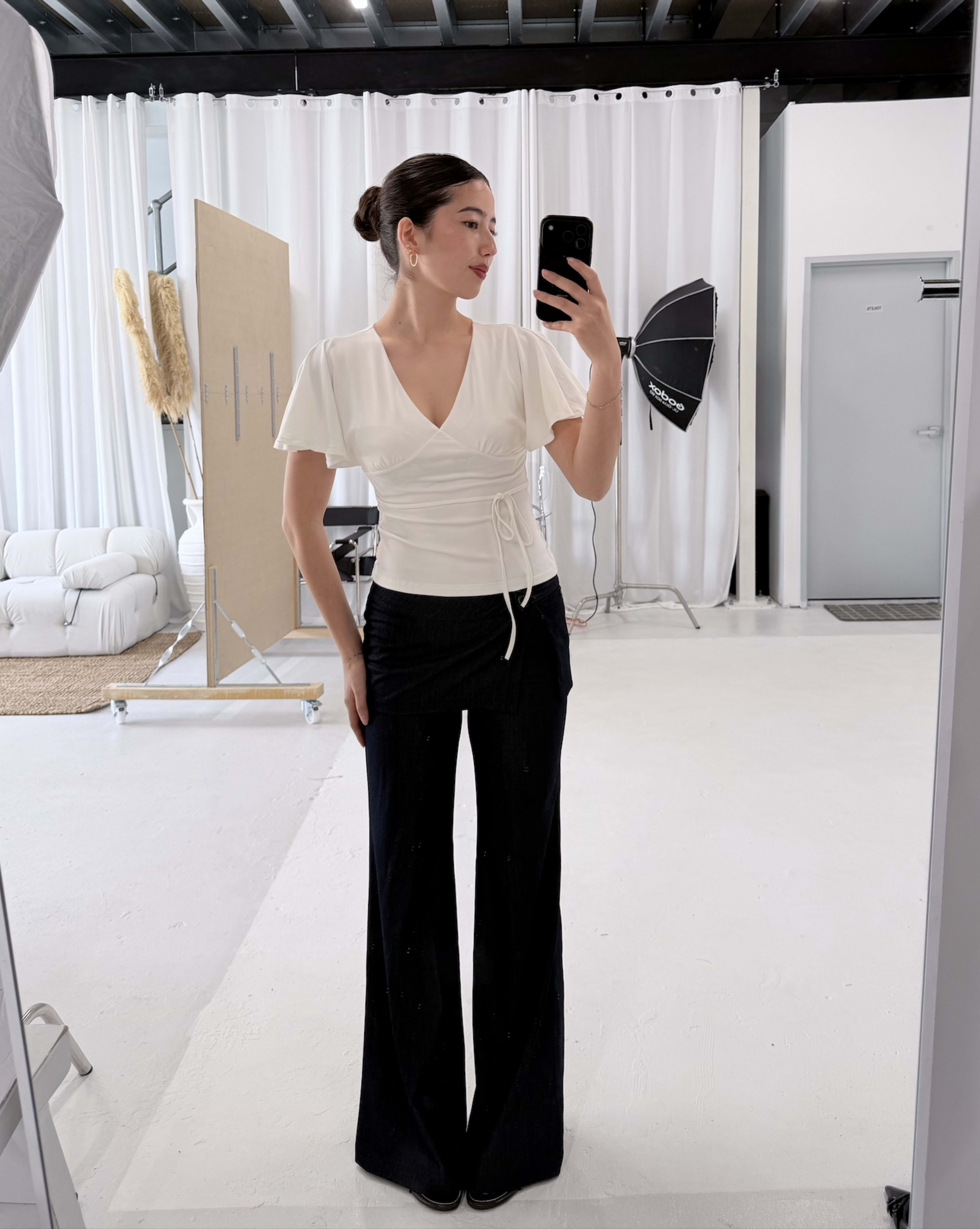 A woman stands in front of a mirror wearing a form-fitting white top and black flared pants in a well-lit studio space.