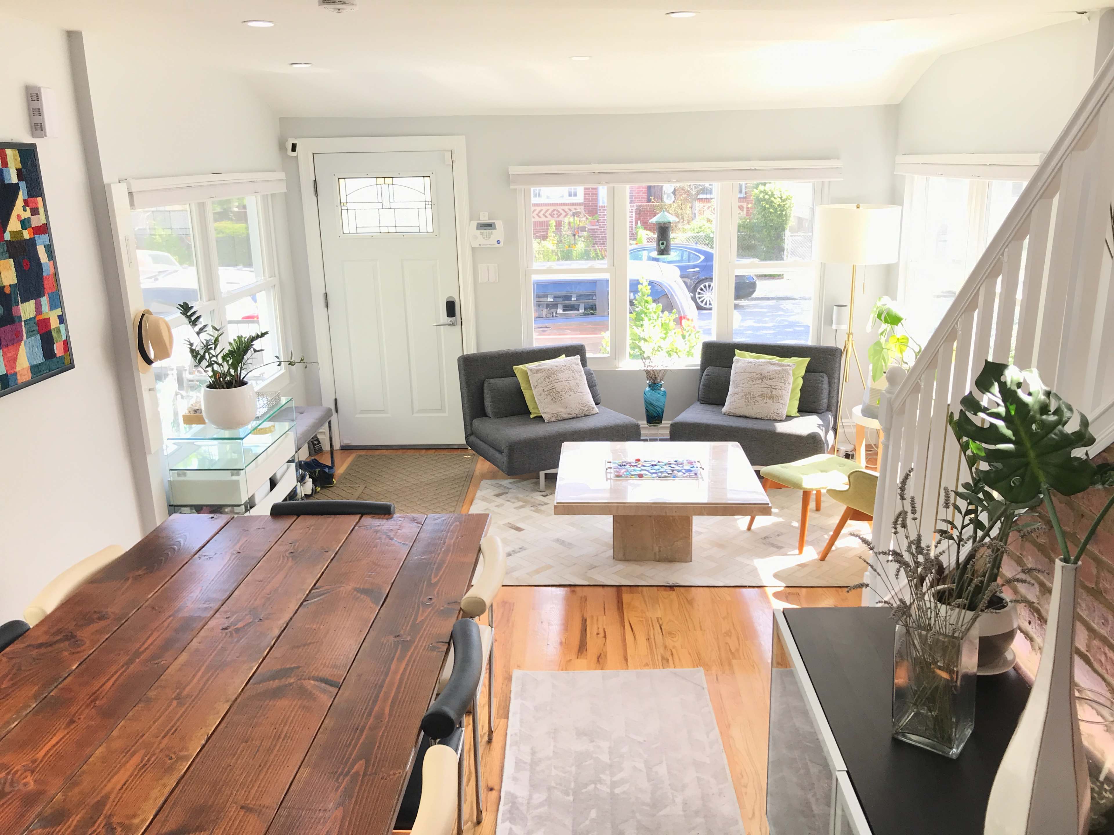 The image shows a bright living space featuring a gray sofa, a coffee table, a dining area with a wooden table, and large windows that allow natural light to enter.